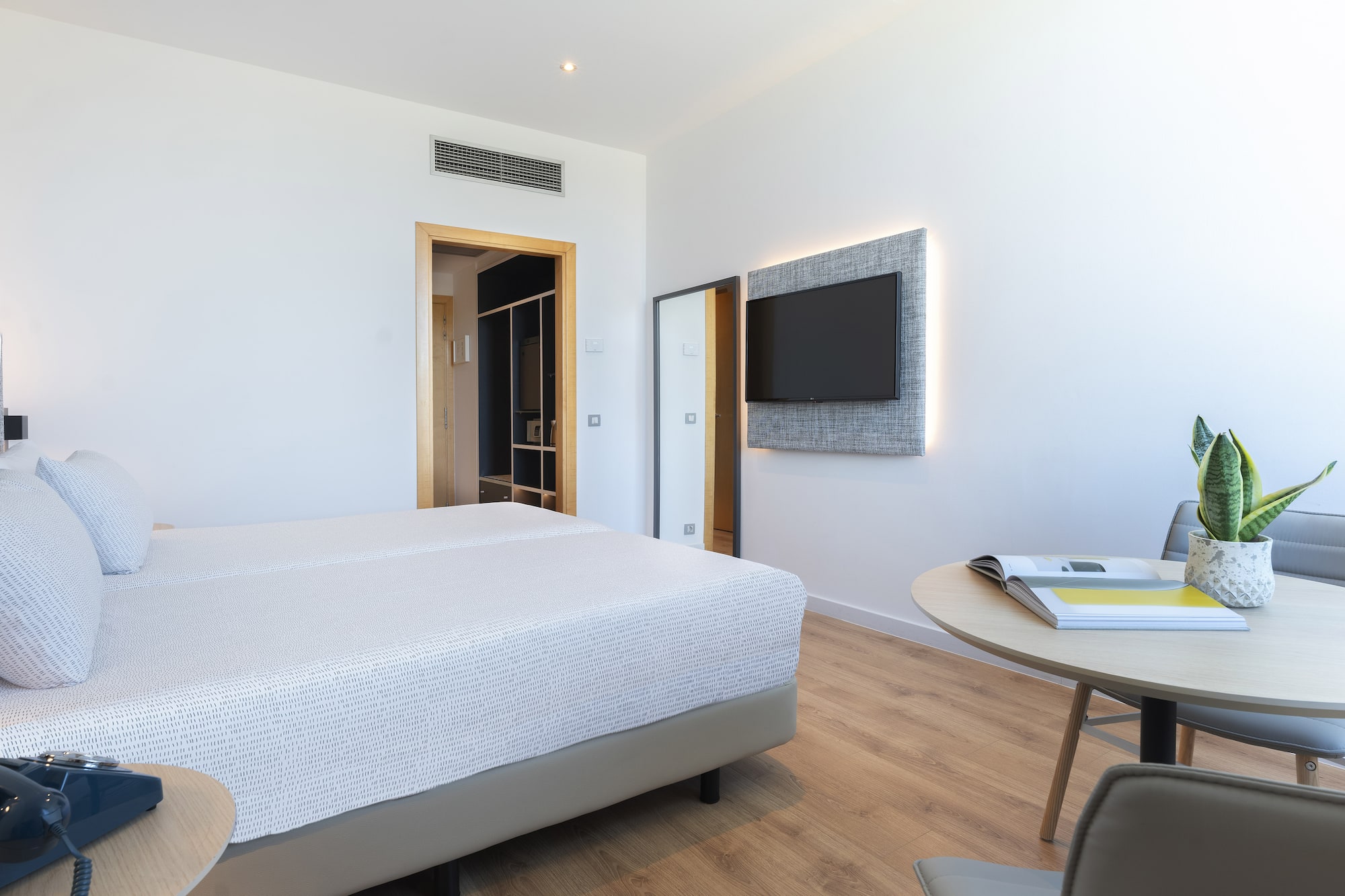 Modern hotel room with twin beds, wall-mounted TV, wooden floor, and round table.