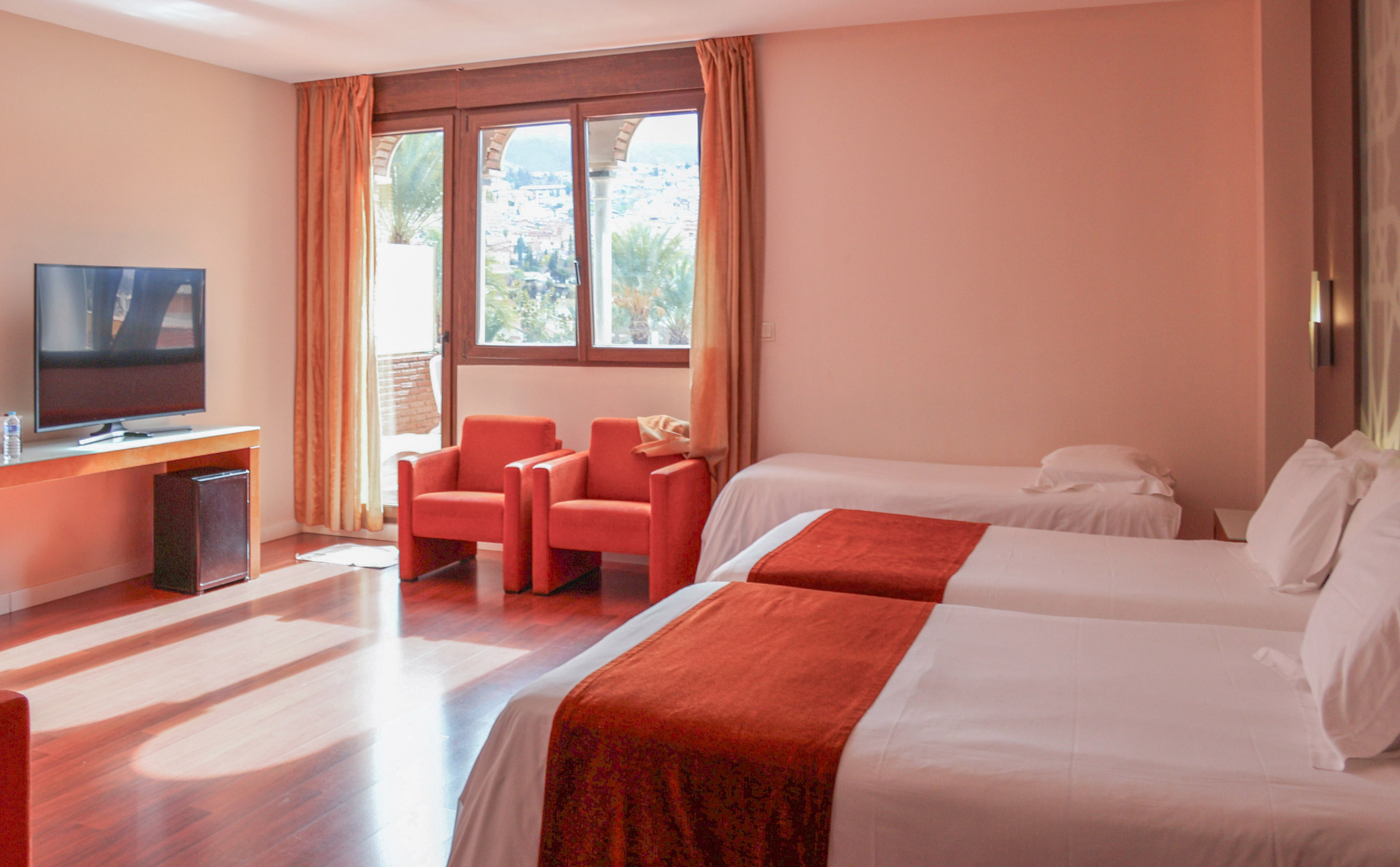 Spacious hotel triple room with three beds, armchairs, TV, and city view.
