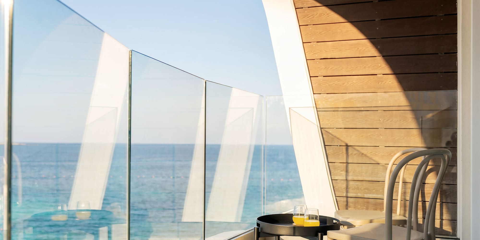 Sunny hotel balcony with sea view, glass railing, table, juice, and modern chairs.
