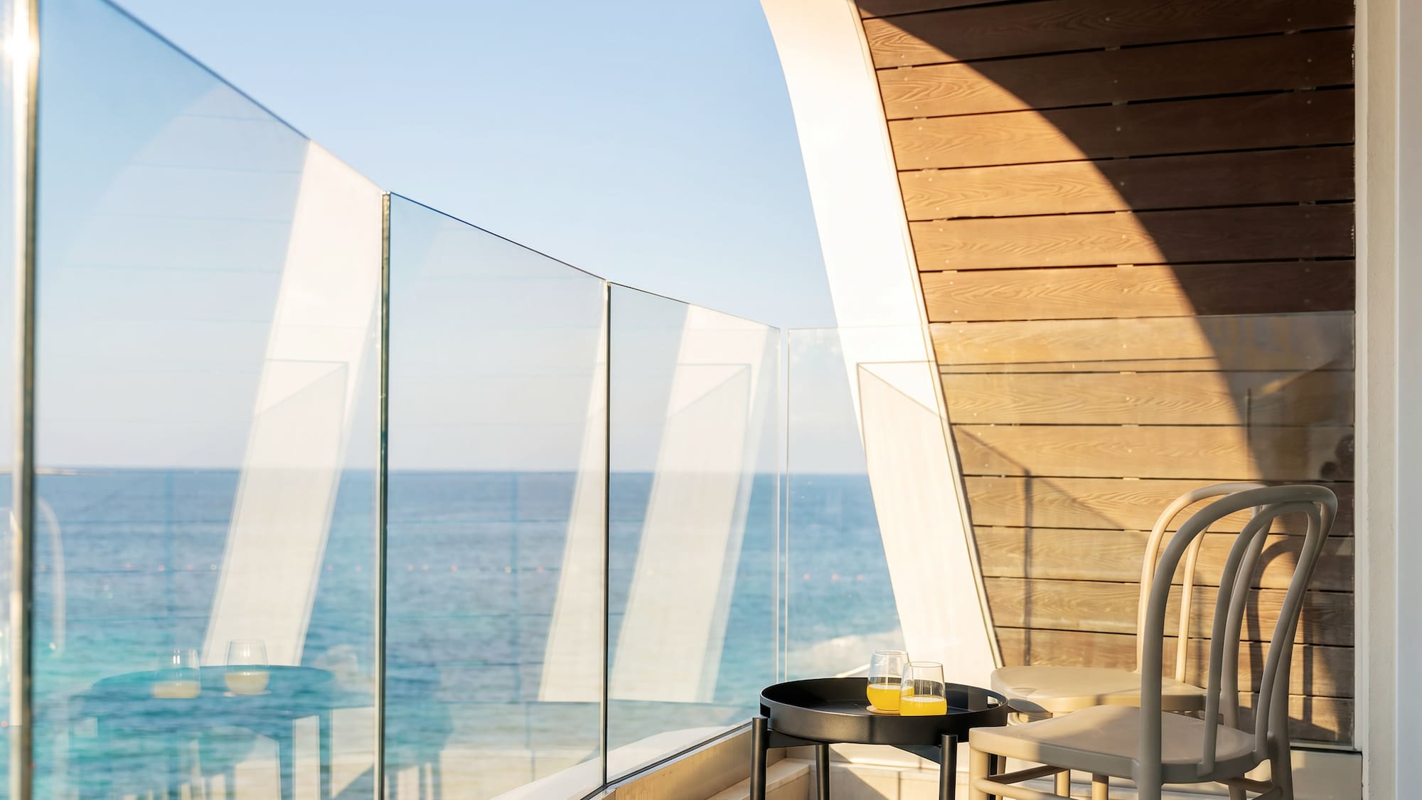 Sunny hotel balcony with sea view, glass railing, table, juice, and modern chairs.