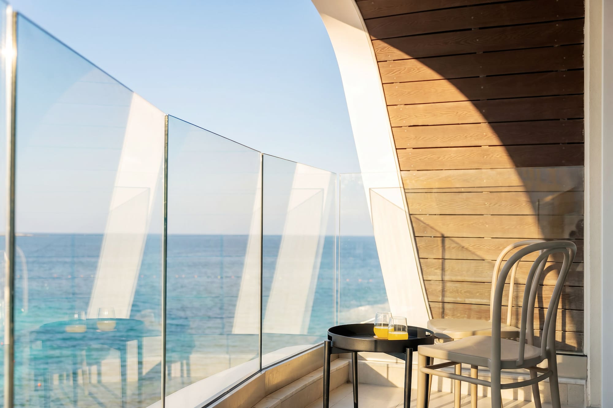 Sunny hotel balcony with sea view, glass railing, table, juice, and modern chairs.