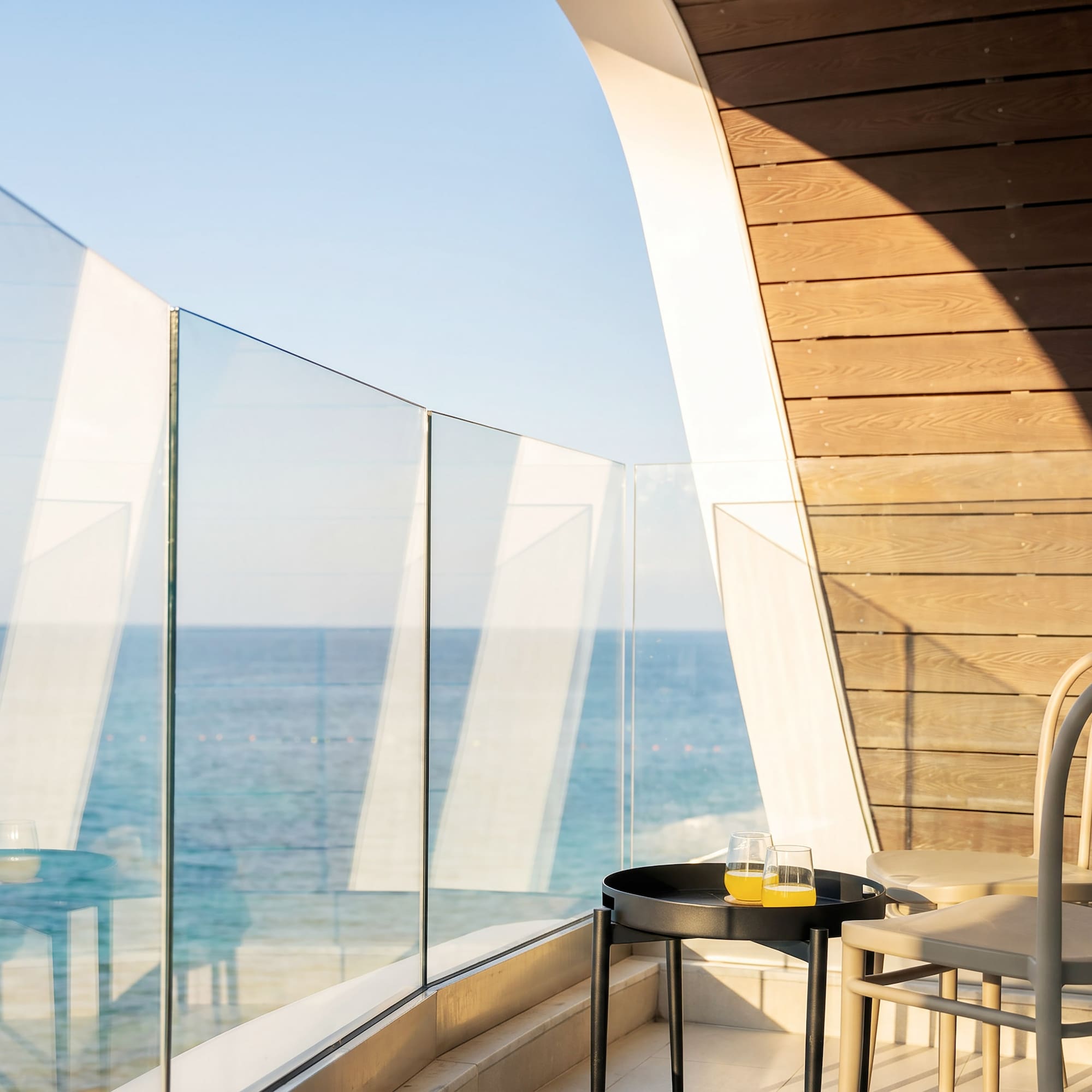 Sunny hotel balcony with sea view, glass railing, table, juice, and modern chairs.