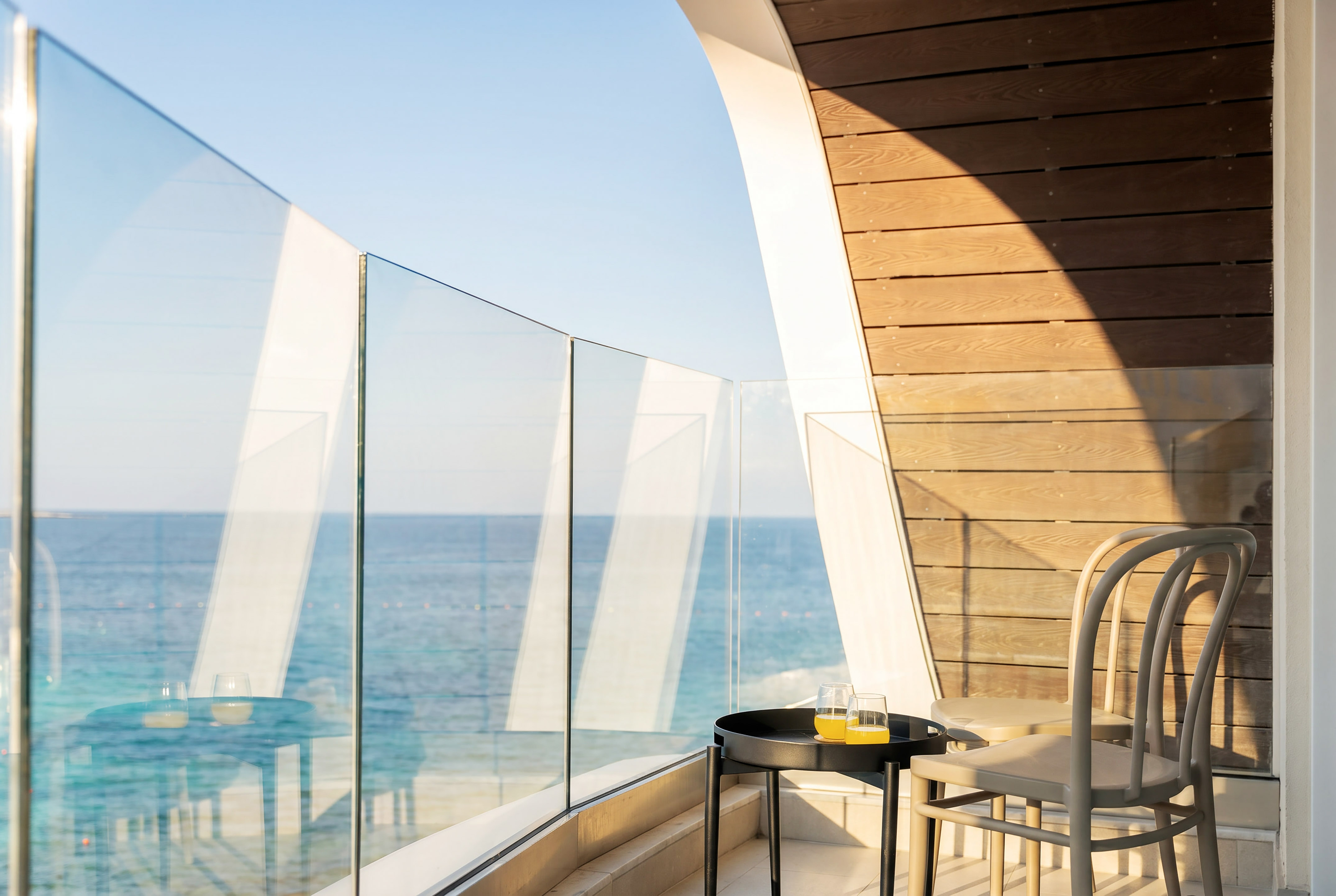 Sunny hotel balcony with sea view, glass railing, table, juice, and modern chairs.
