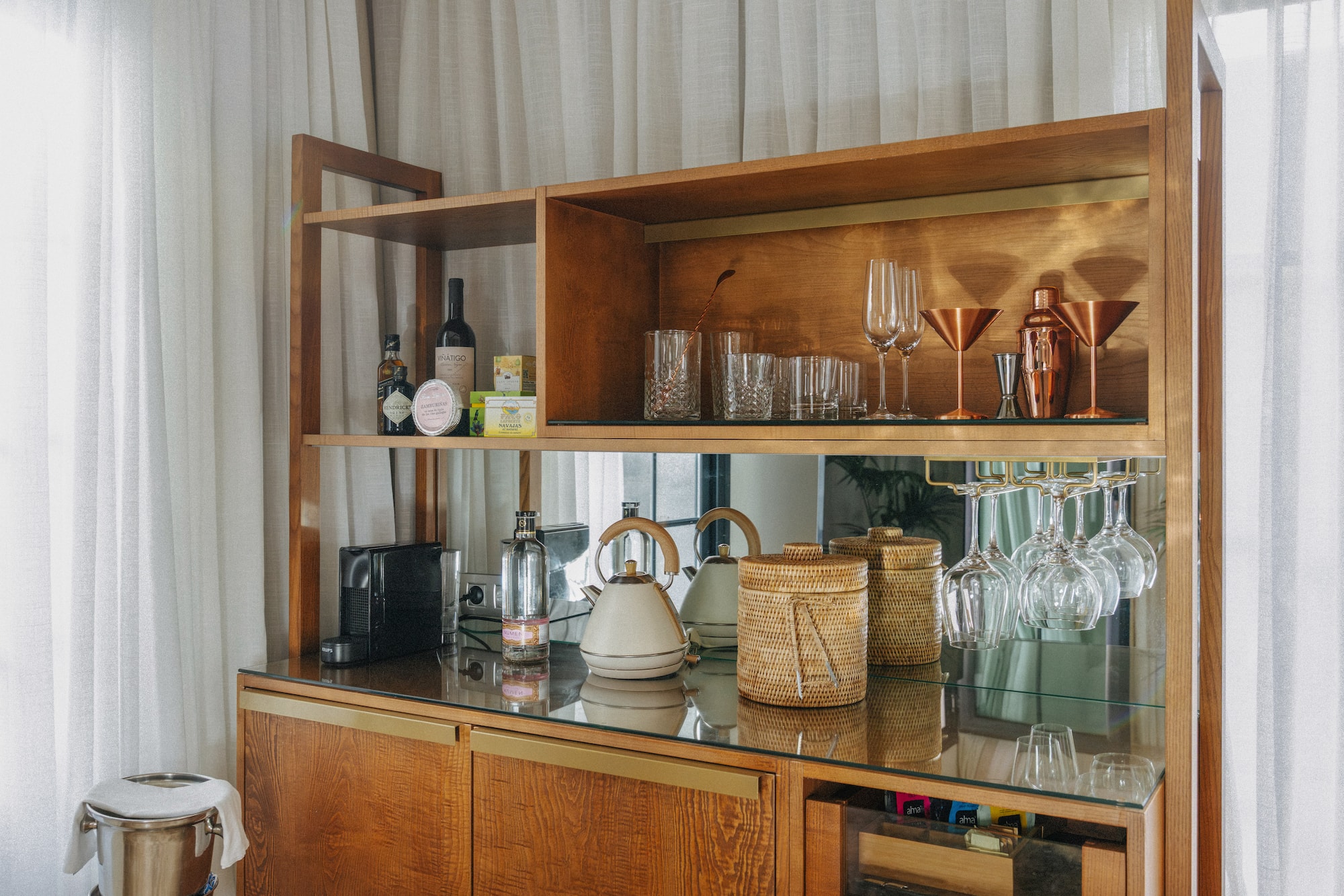 Wooden minibar cabinet with bottles, cocktail glasses, Nespresso, and kettle.