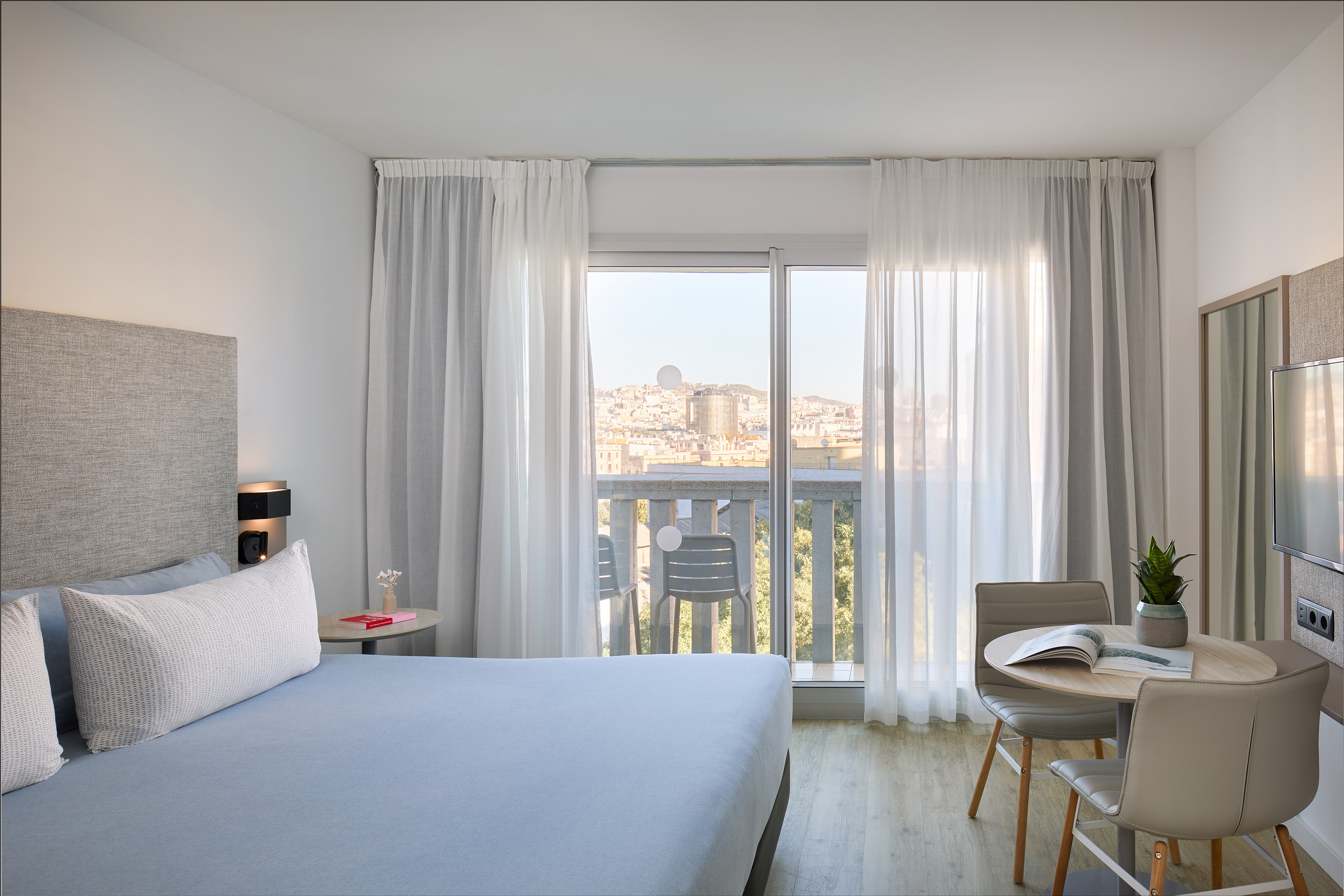 Modern hotel room with bed, city view balcony, and dining area.