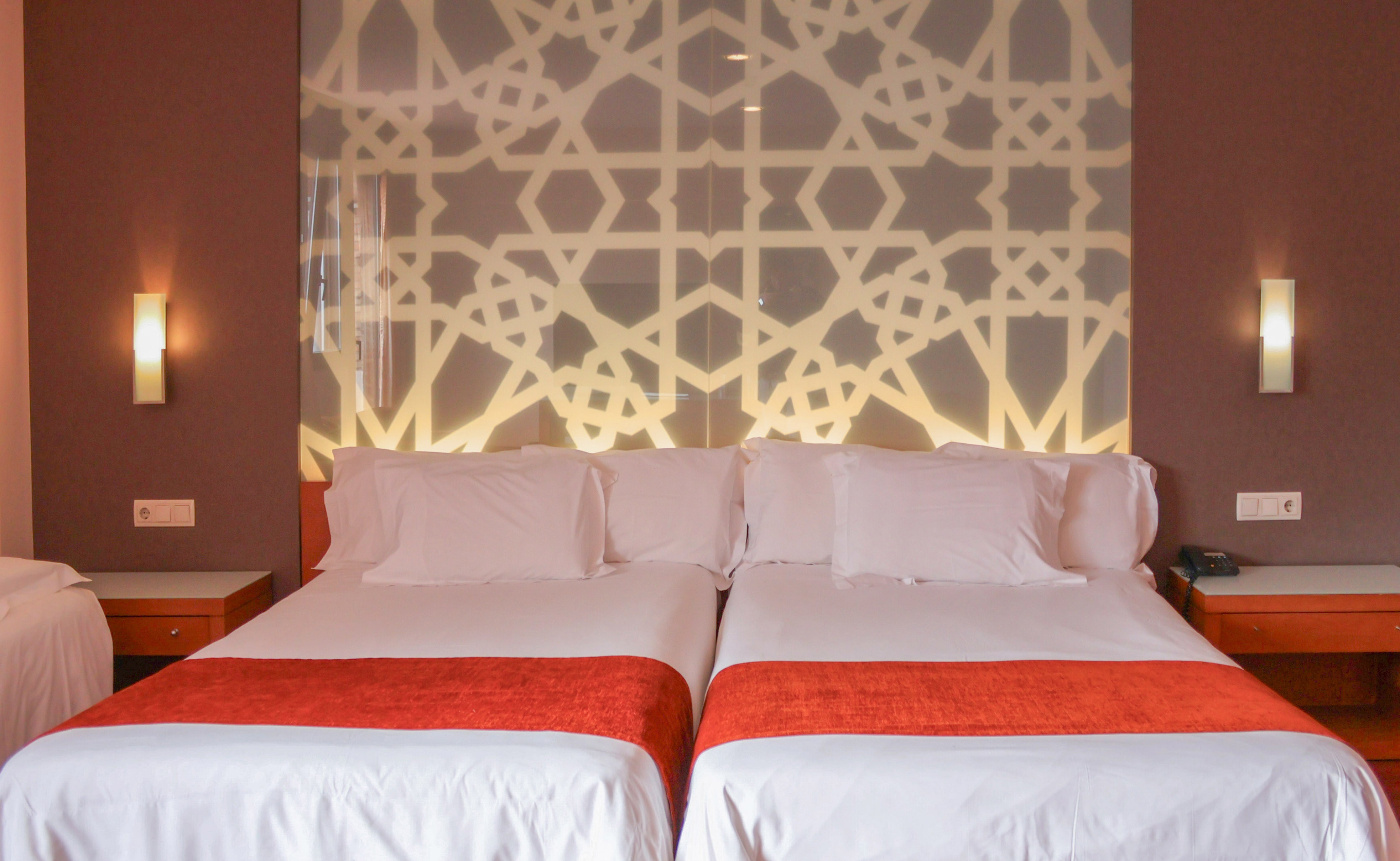 Modern hotel room with twin beds, orange throws, and backlit Moroccan geometric headboard.