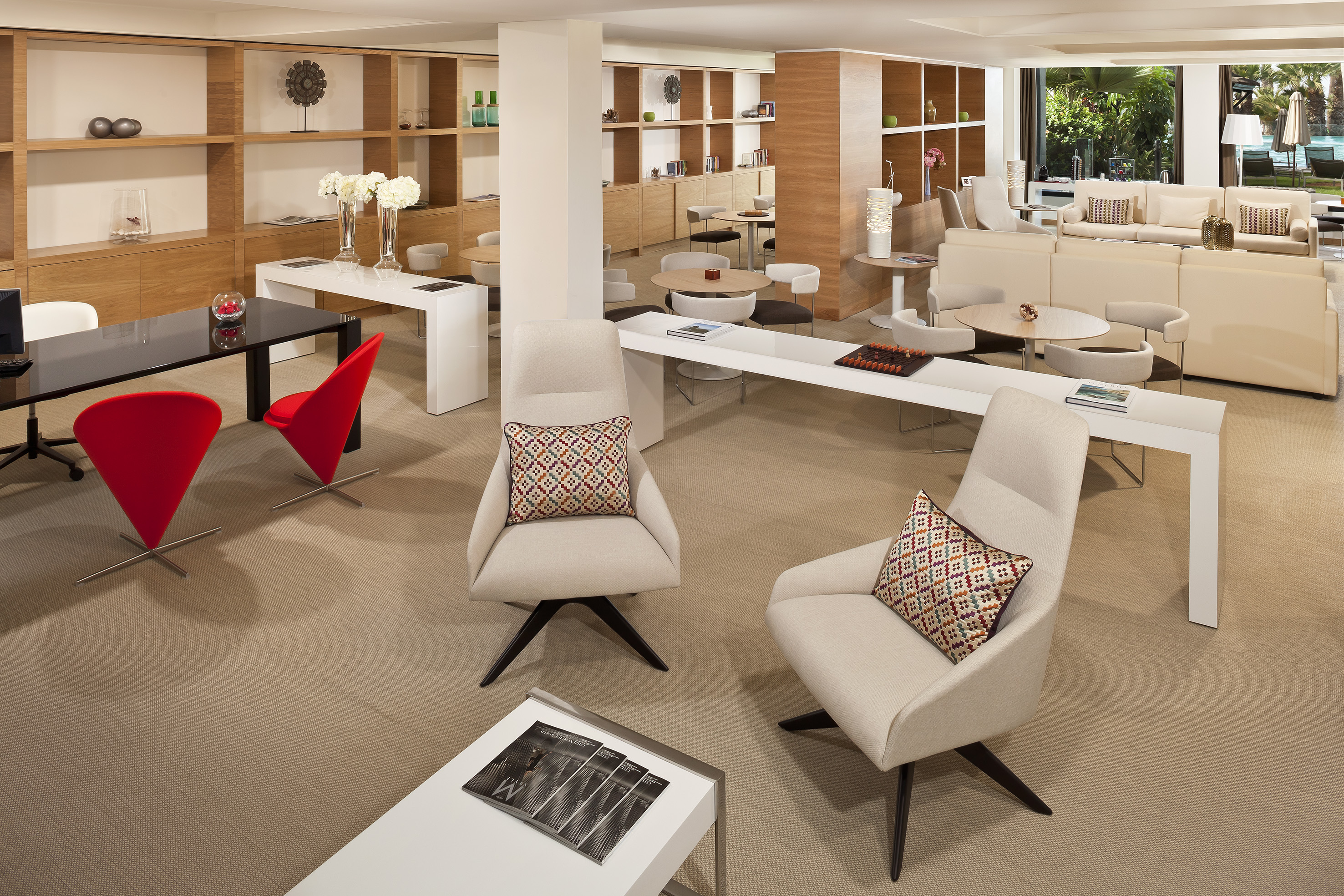 Modern lounge with red chairs, beige sofas, wooden shelves, and outdoor view.