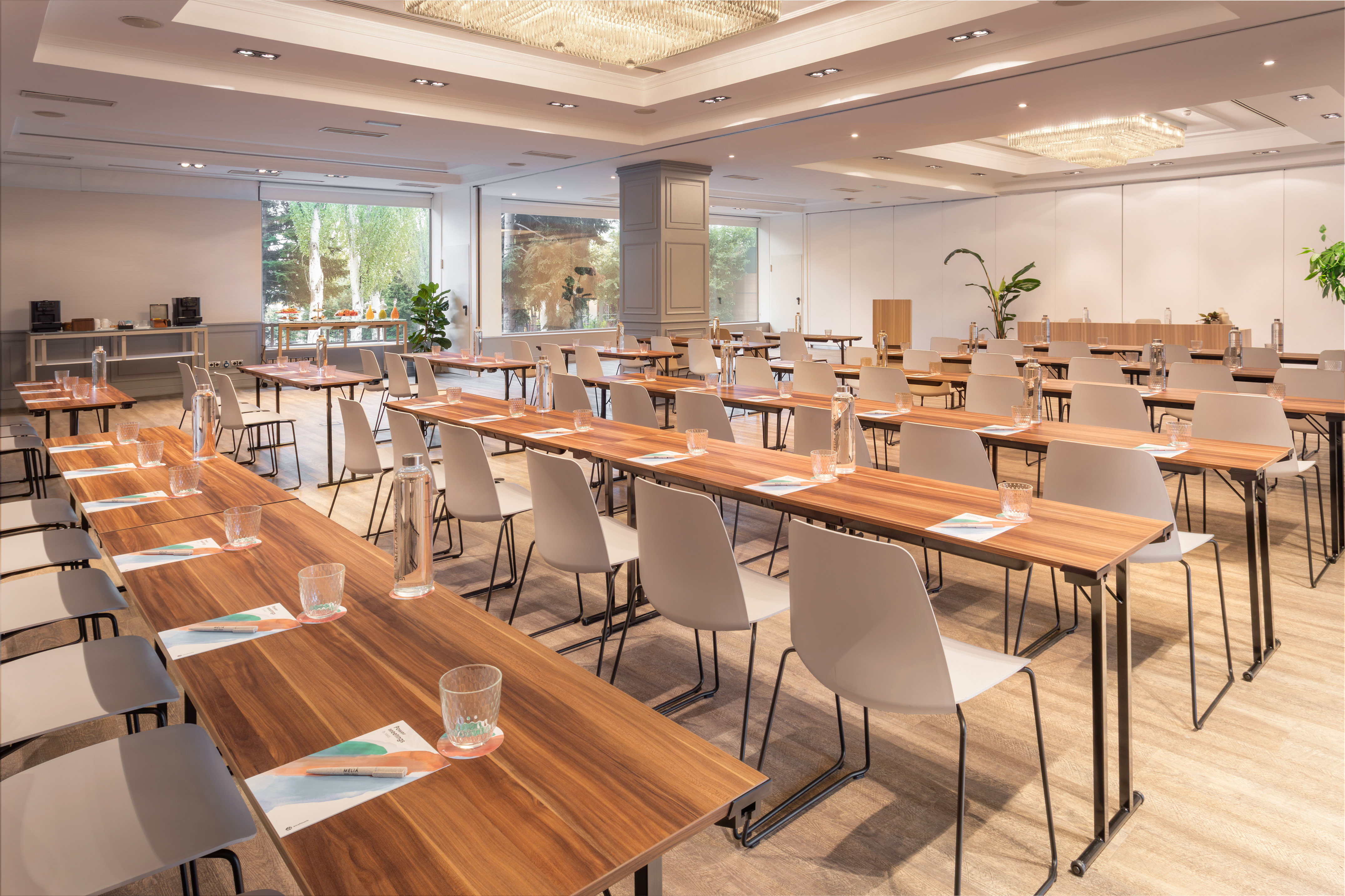 Bright conference room with rows of tables, chairs, and large windows overlooking greenery.