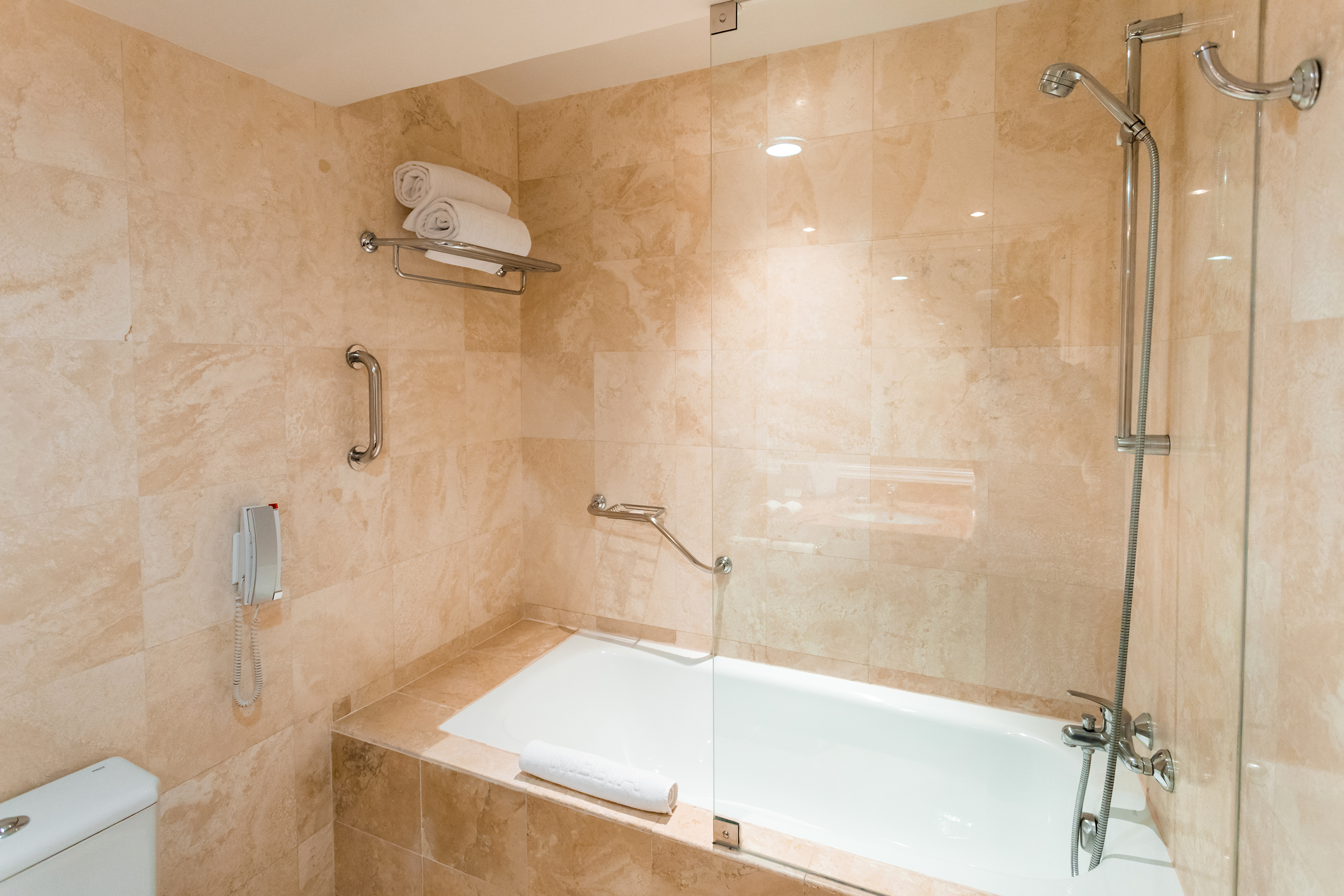 Accessible hotel bathroom with beige tiles, bathtub, shower, and grab bars.