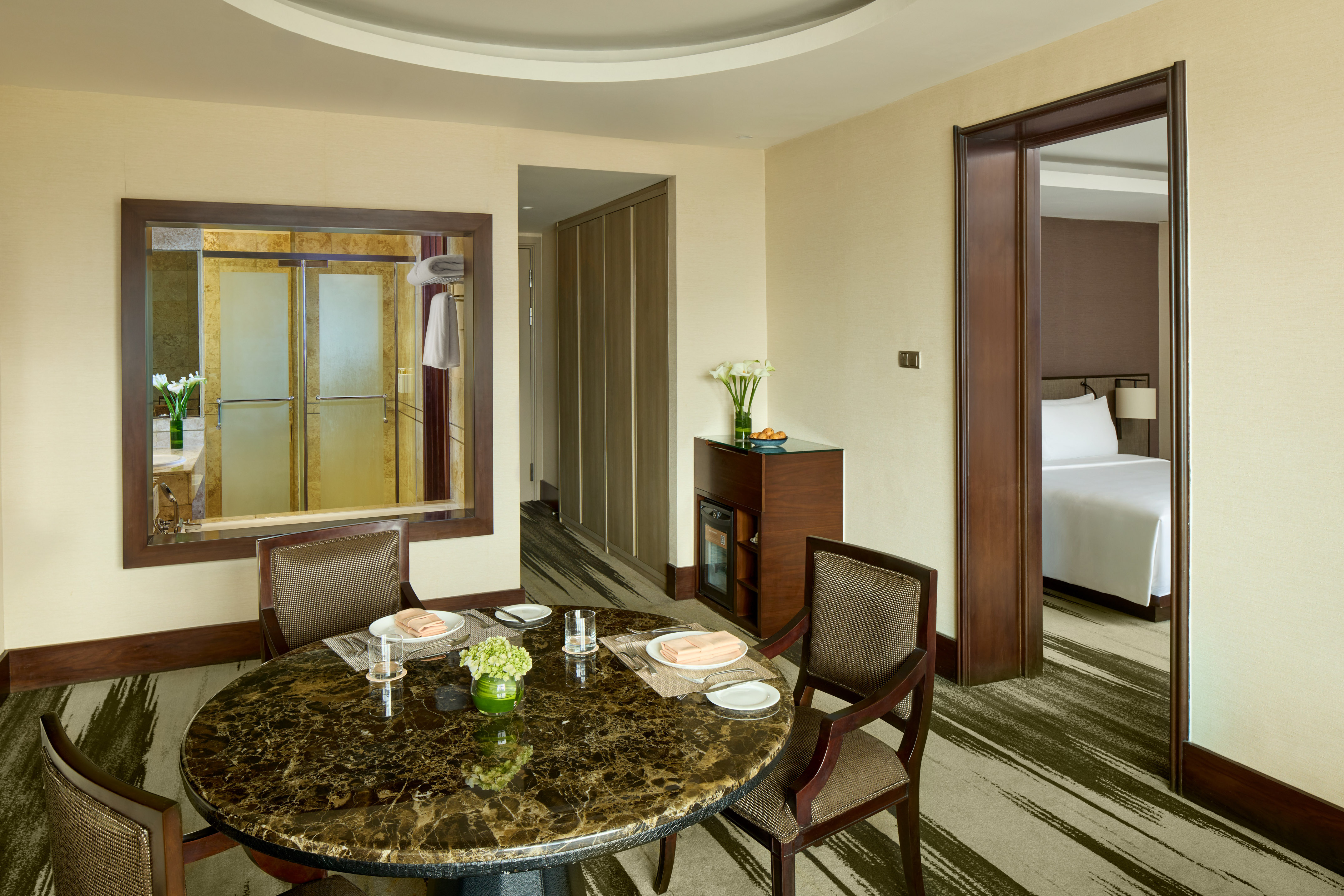 Hotel suite dining area with marble table, reflected bathroom, and bedroom.