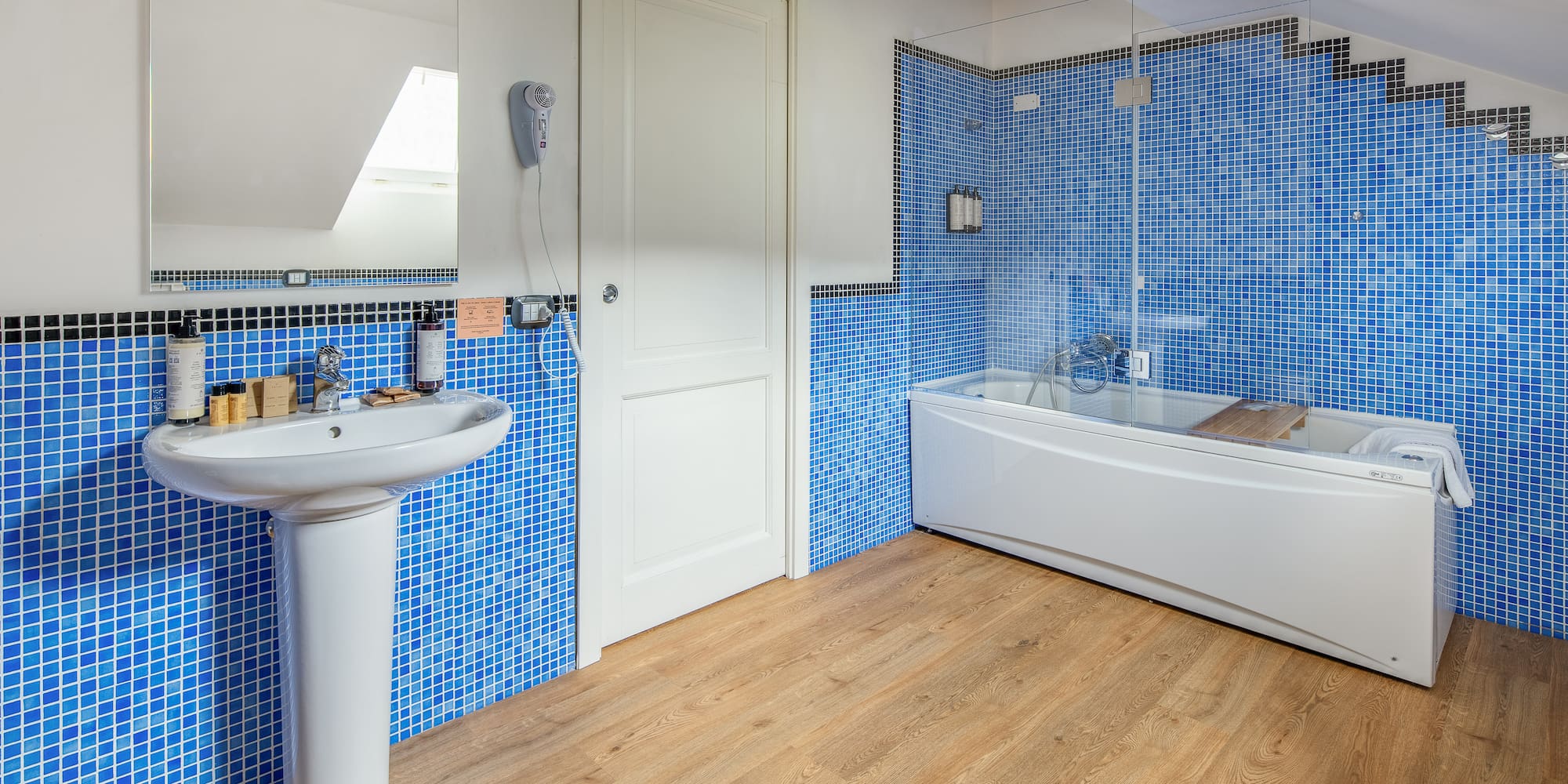 Blue mosaic tiled bathroom with white sink, bathtub, and wood floor.