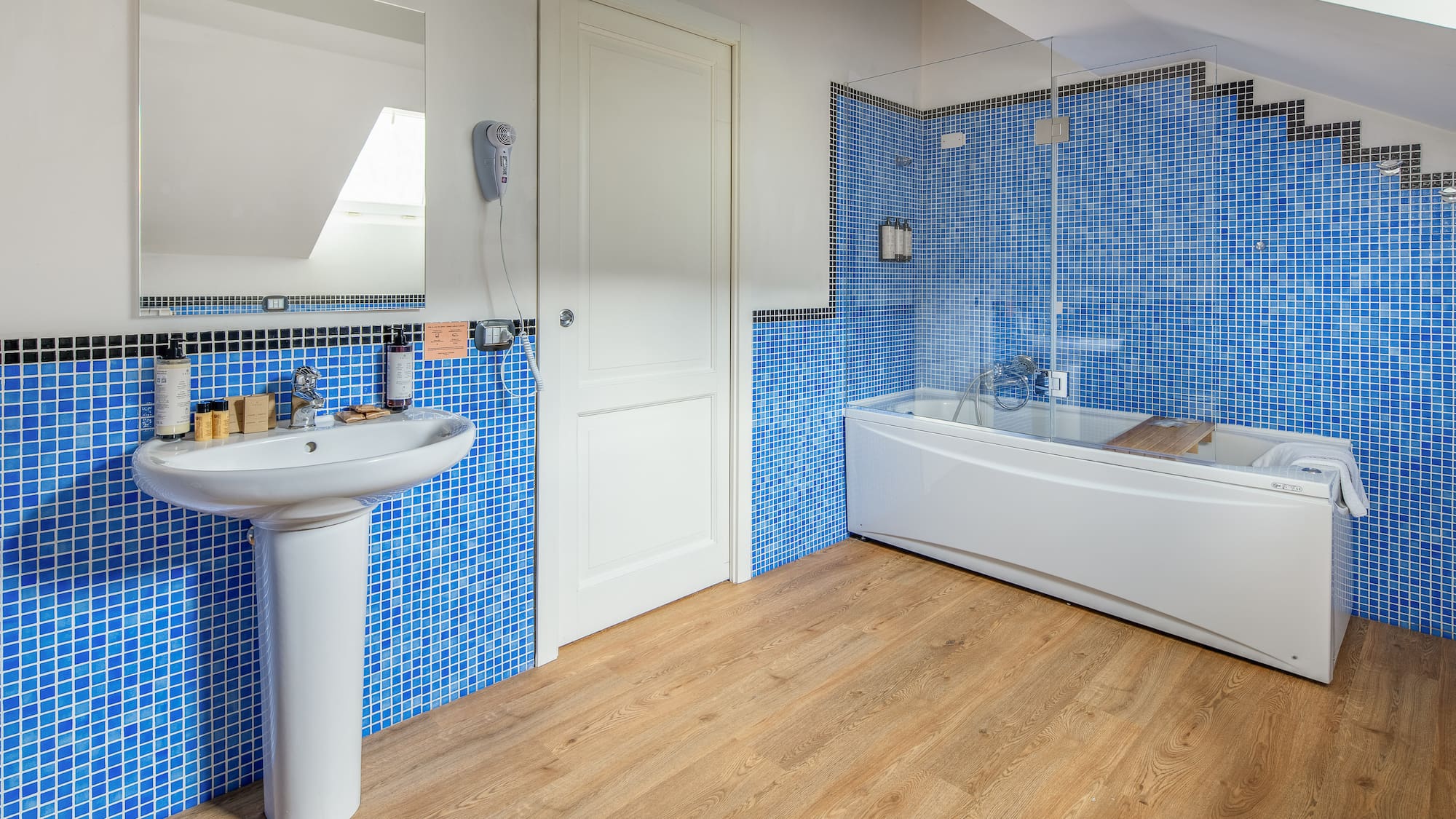 Blue mosaic tiled bathroom with white sink, bathtub, and wood floor.