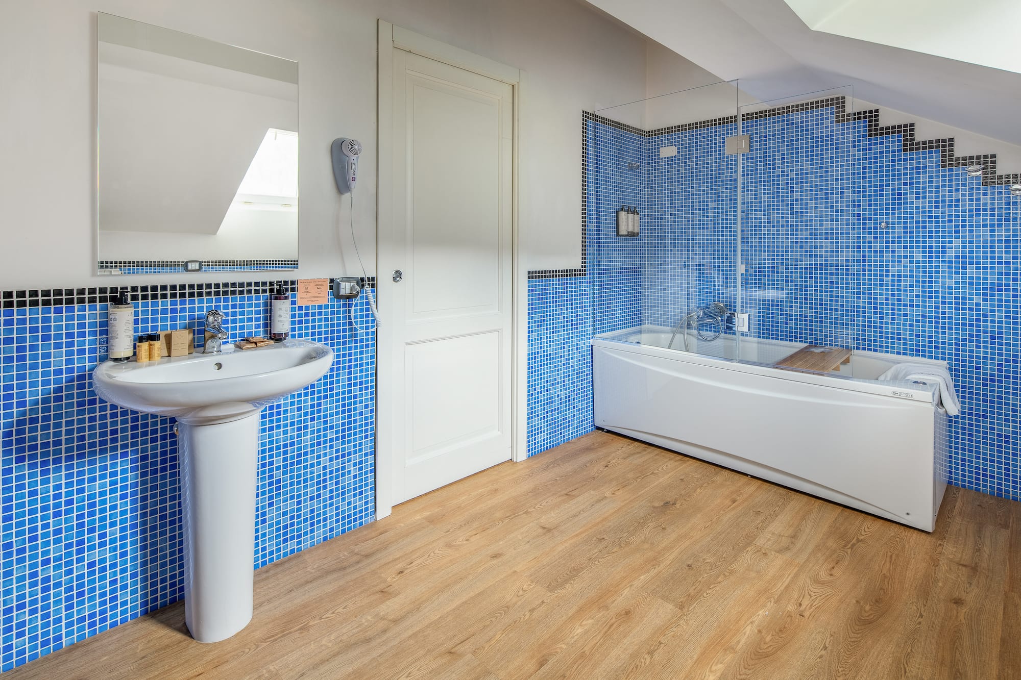 Blue mosaic tiled bathroom with white sink, bathtub, and wood floor.