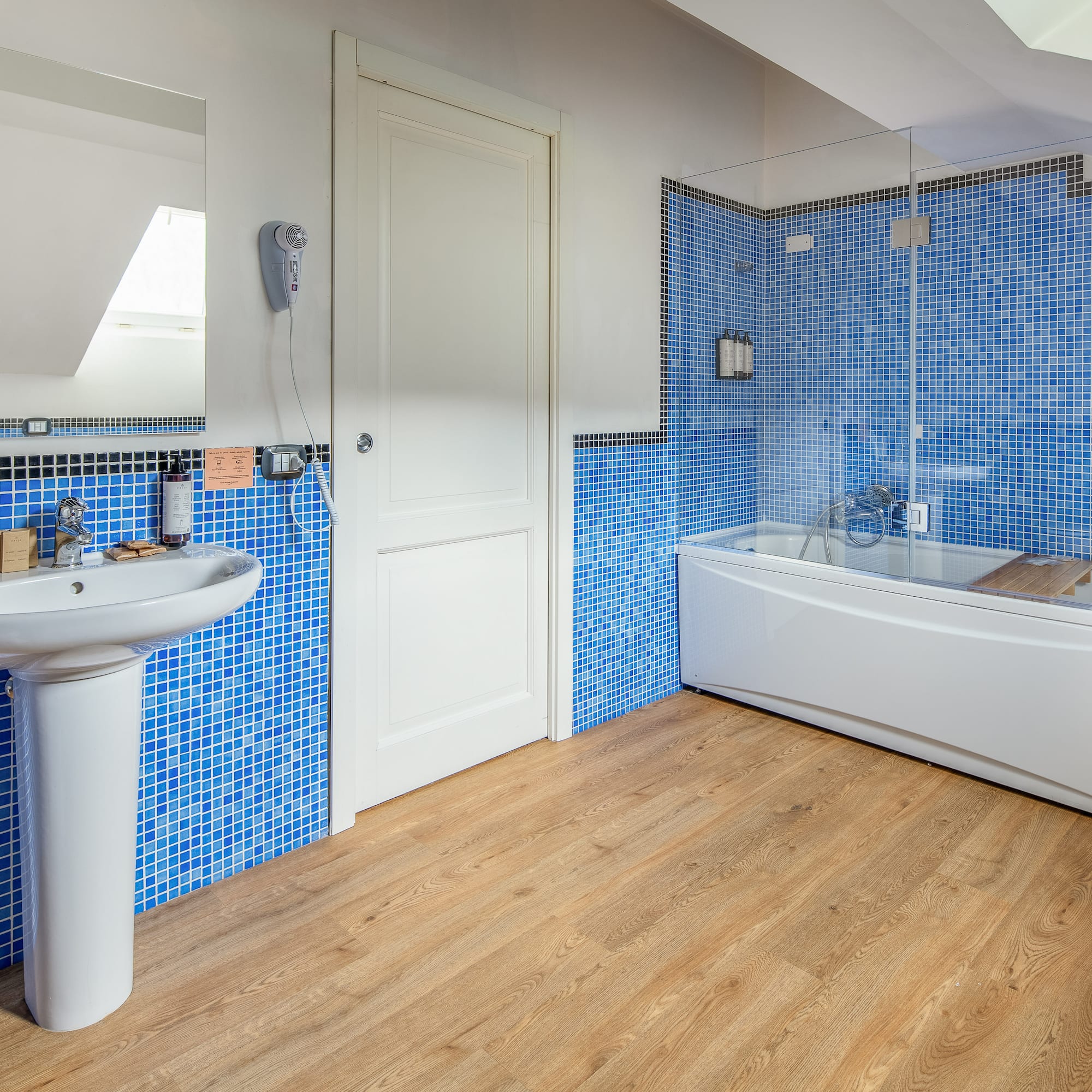 Blue mosaic tiled bathroom with white sink, bathtub, and wood floor.