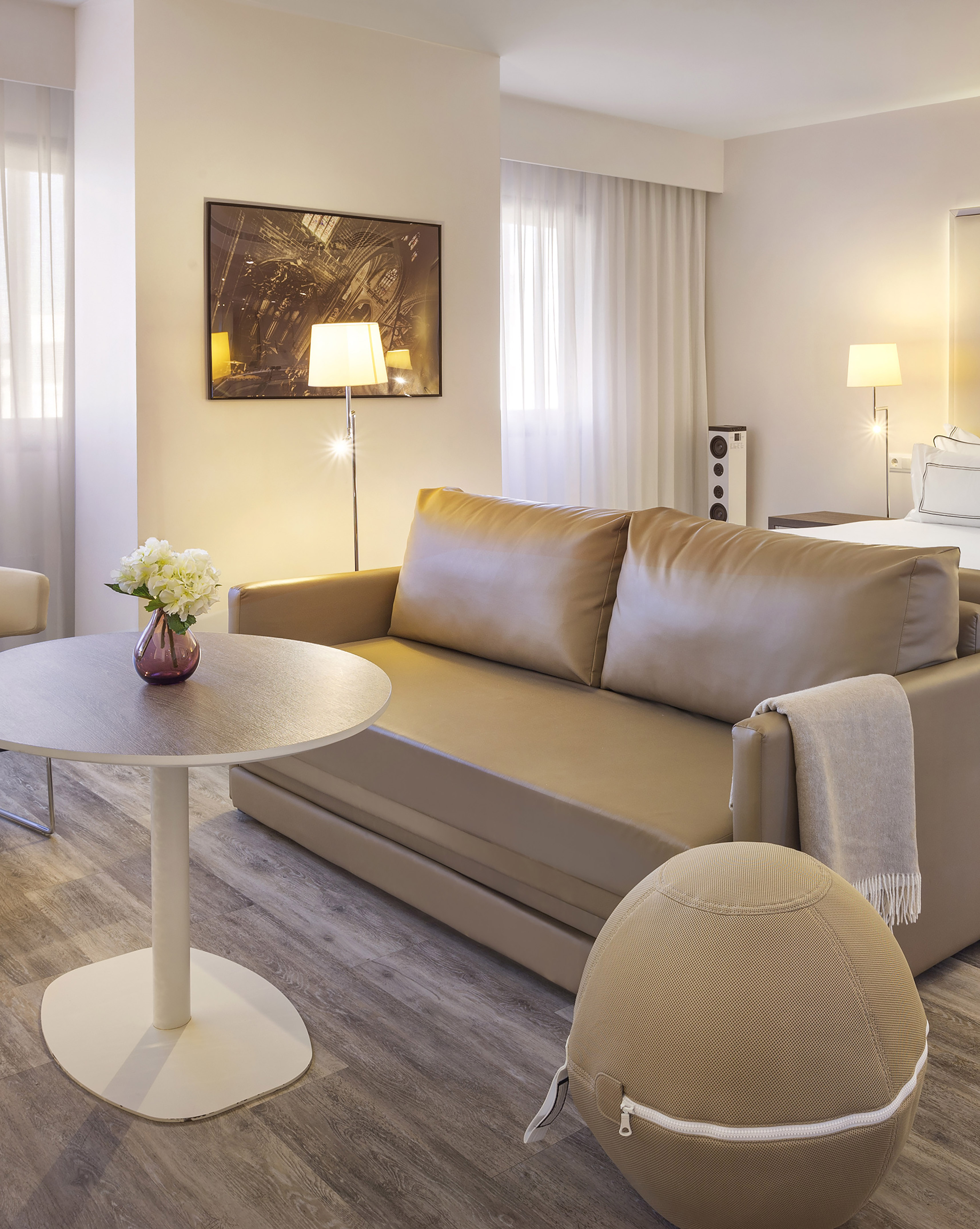 Modern hotel room living area with tan sofa, round table, flowers, and floor lamp.