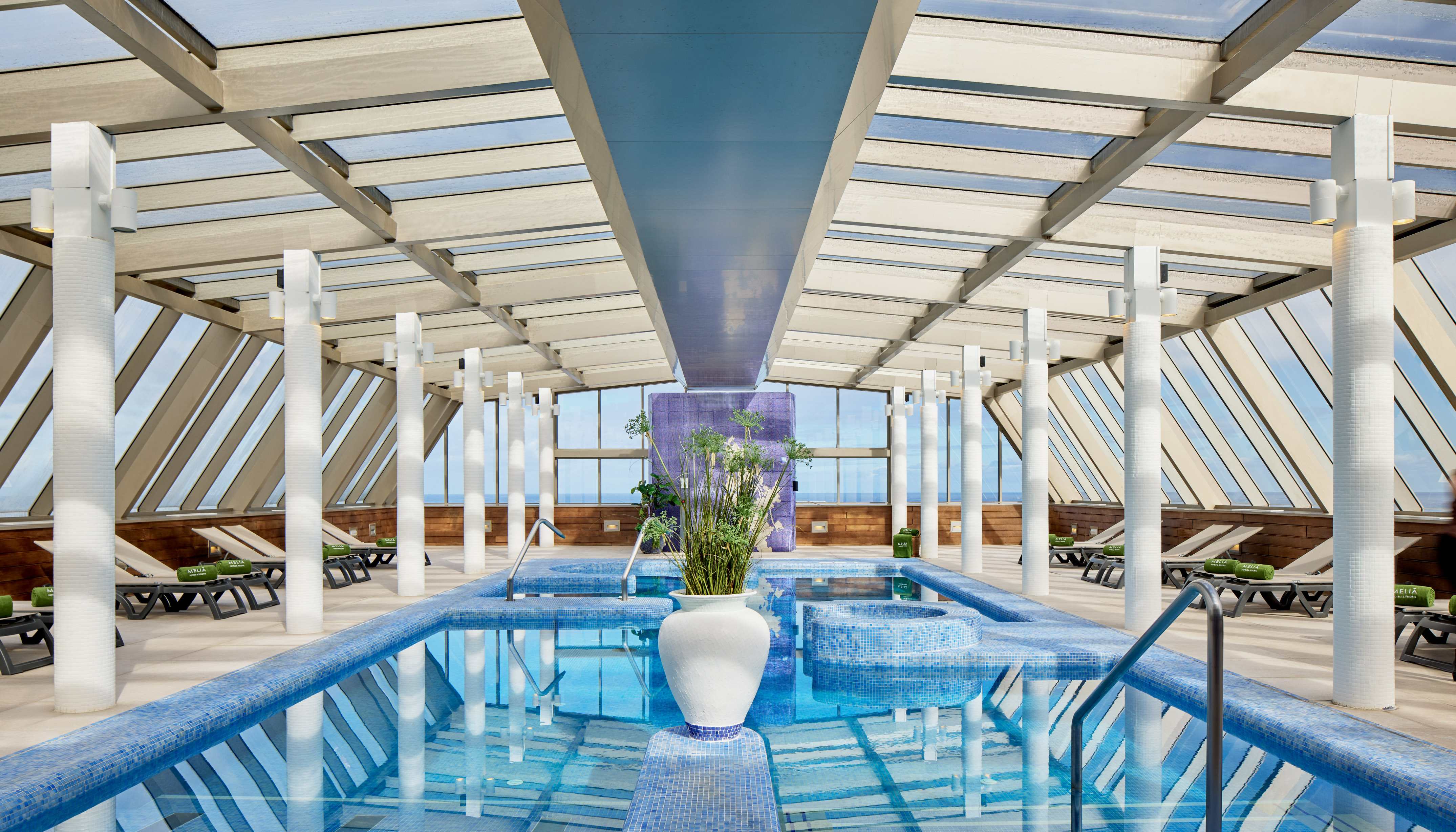 Modern indoor swimming pool with glass roof, ocean view, spa and lounge chairs.