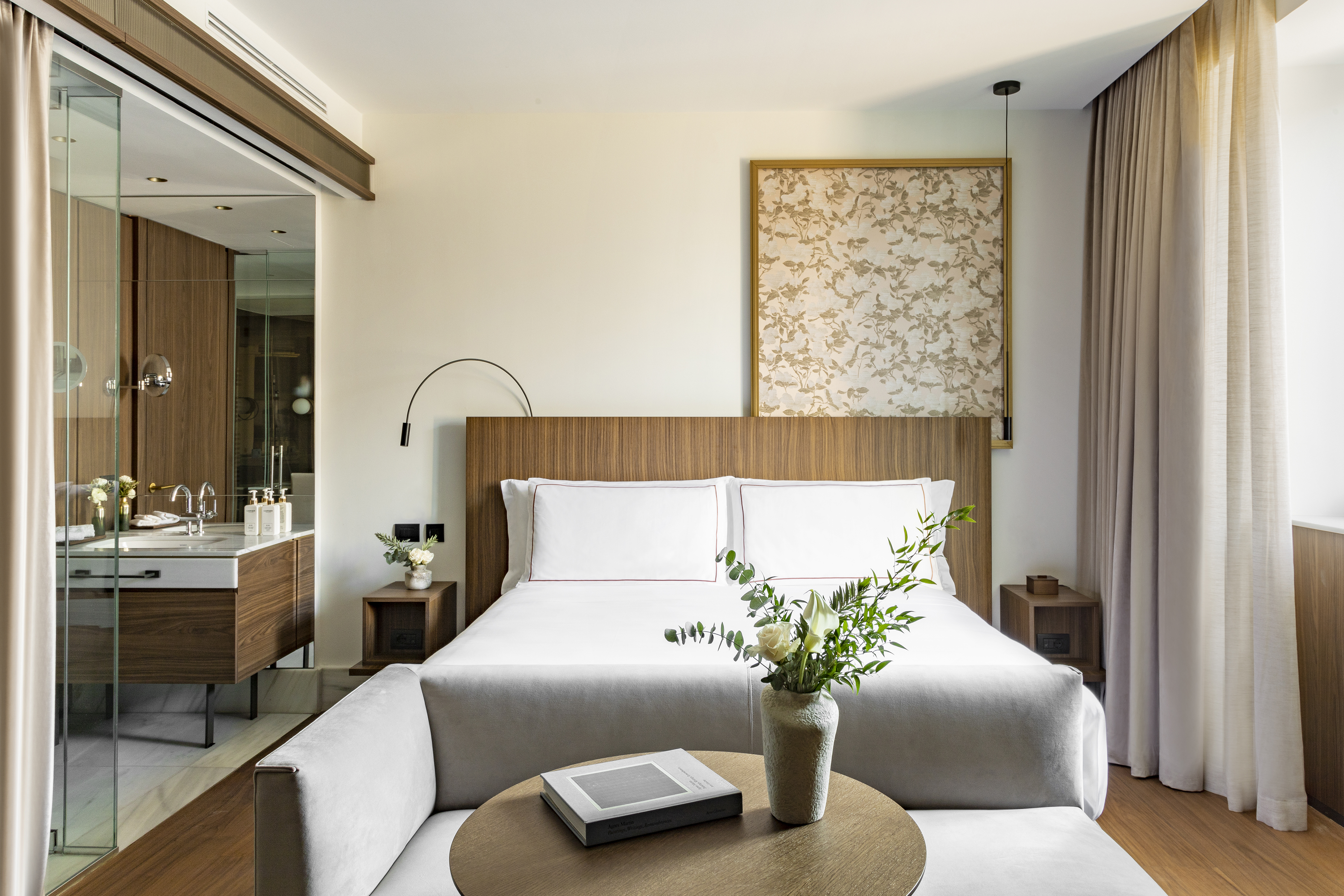 Modern hotel bedroom with white bed, wooden headboard, and glass bathroom.