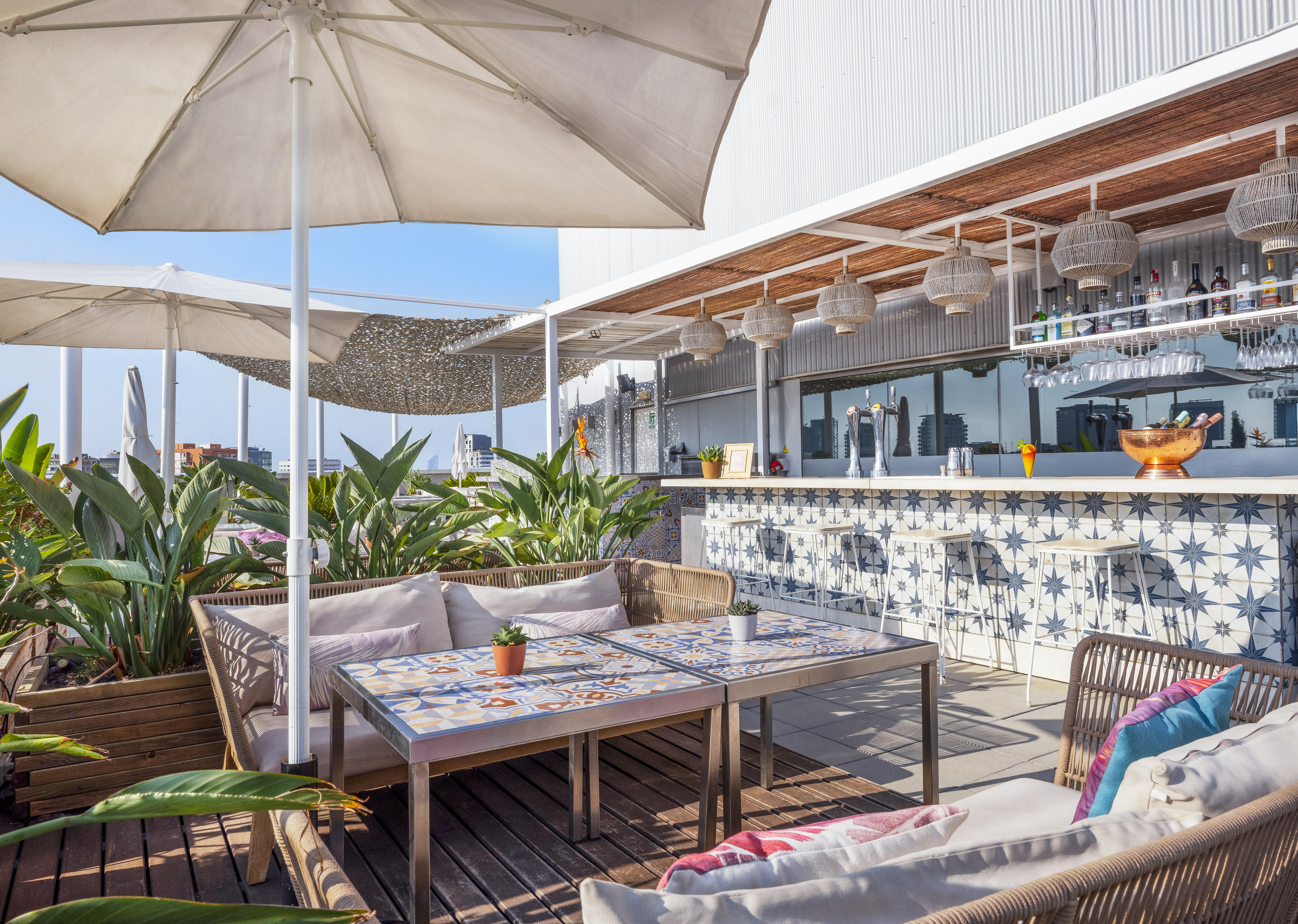 Sunny rooftop bar lounge with tiled tables, green plants, and umbrellas.