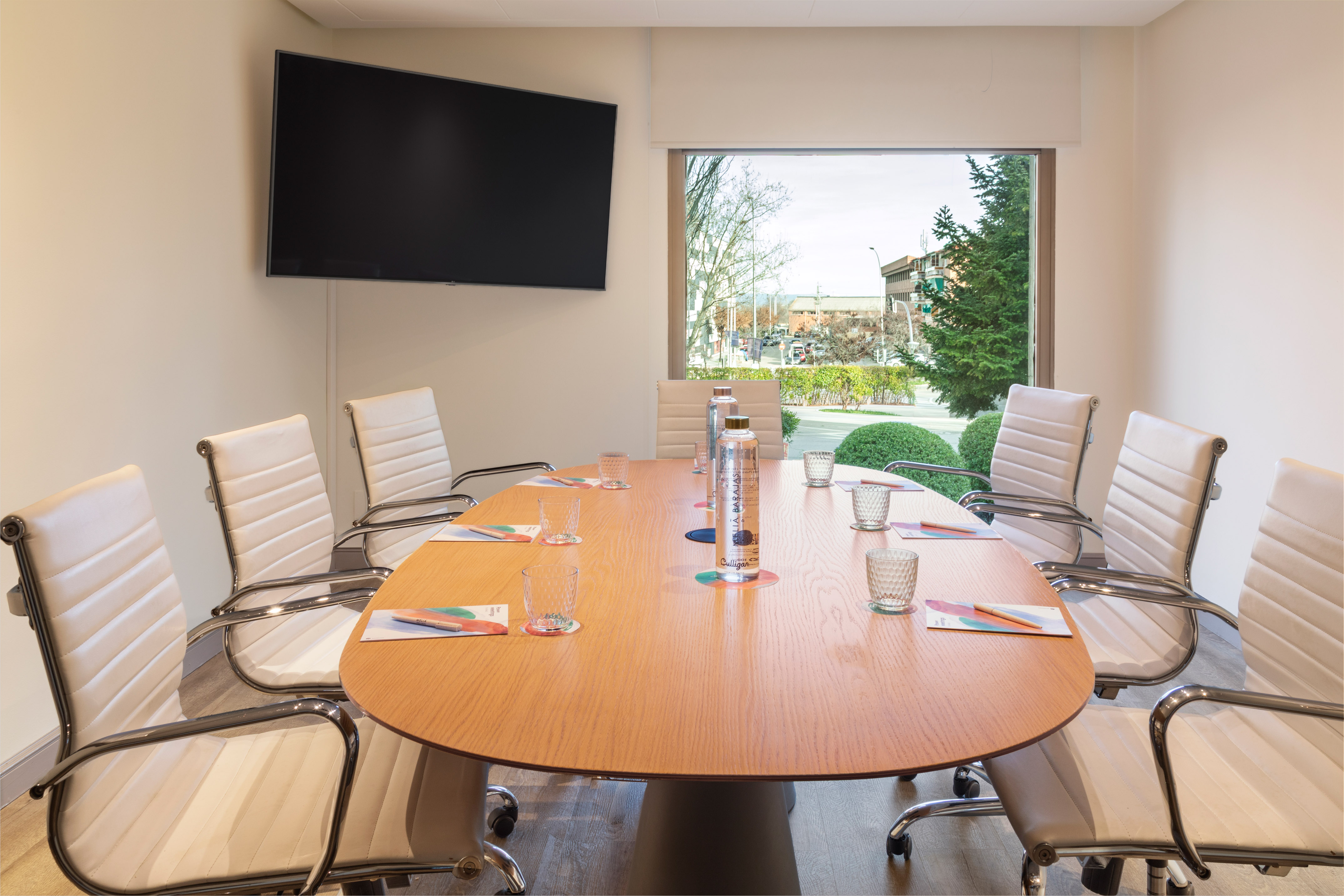 Modern meeting room with oval wooden table, cream chairs, large TV, and window view.