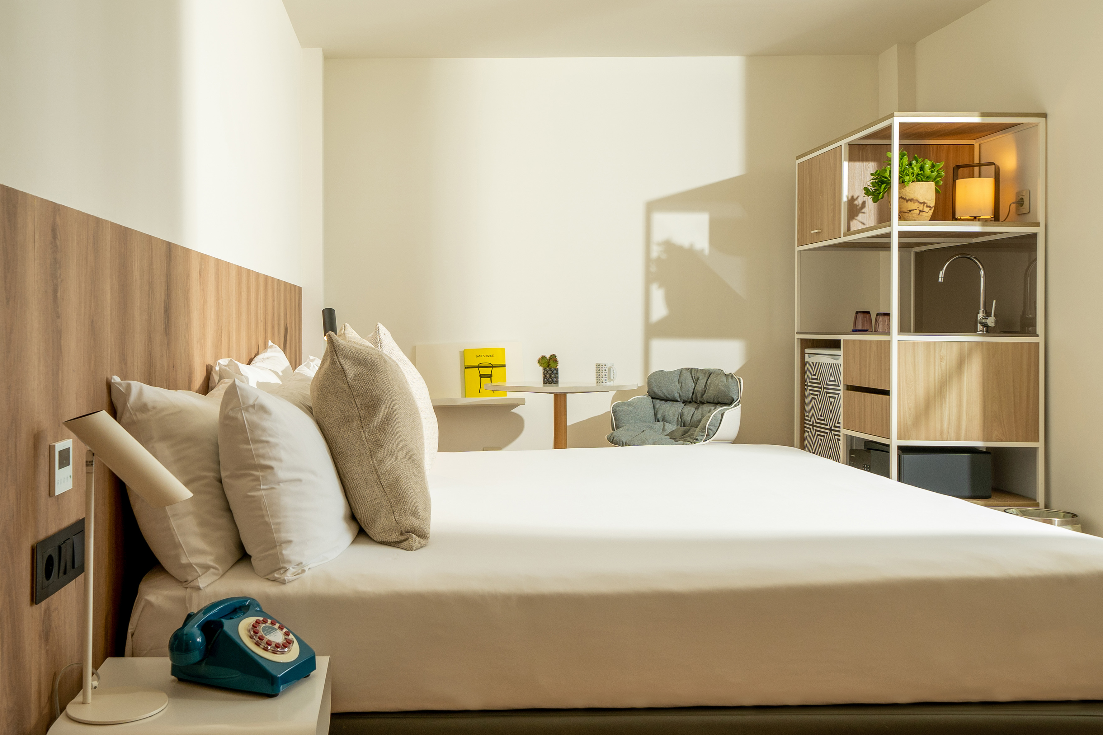 Modern hotel bedroom with white bed, wooden headboard, and compact kitchenette.