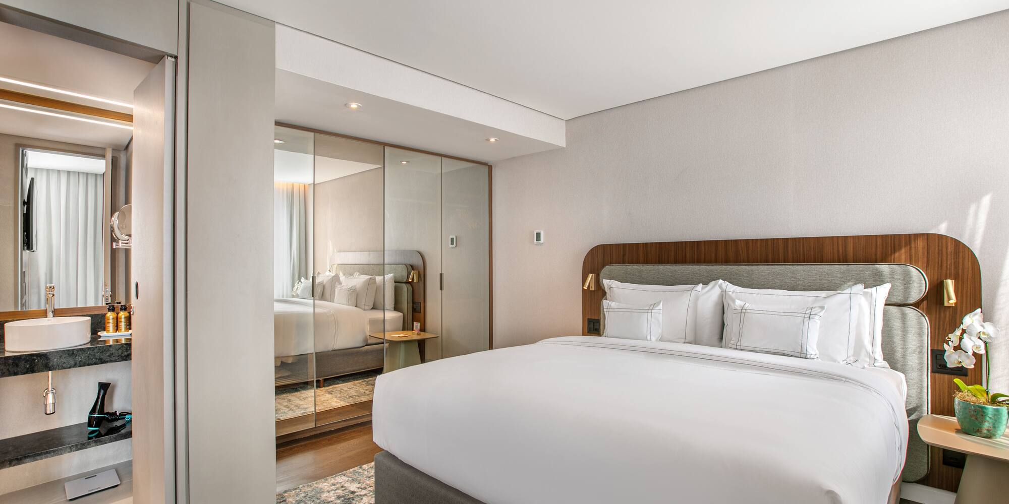 Modern hotel bedroom with king bed, reflective wardrobe, and ensuite vanity.