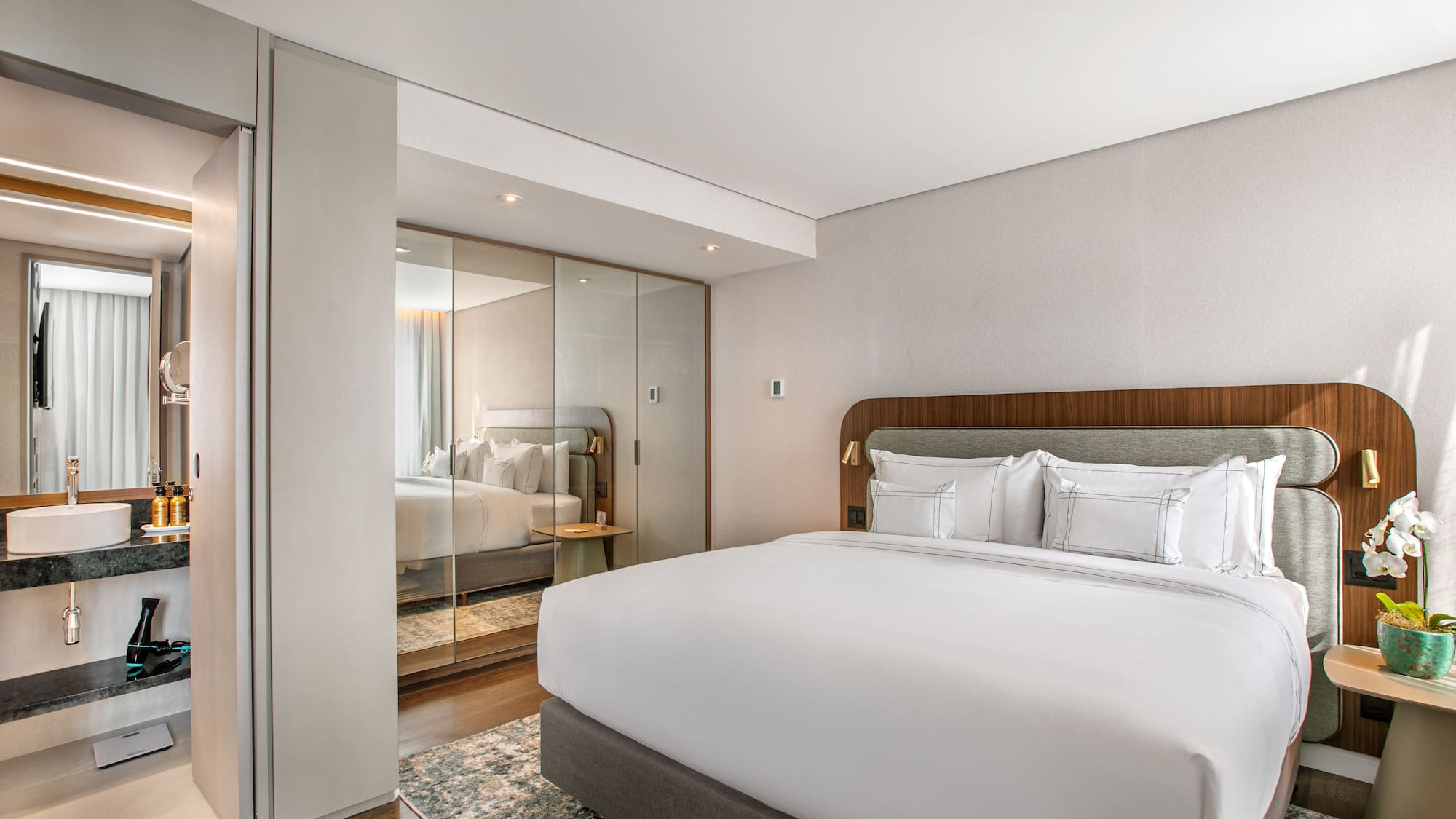 Modern hotel bedroom with king bed, reflective wardrobe, and ensuite vanity.