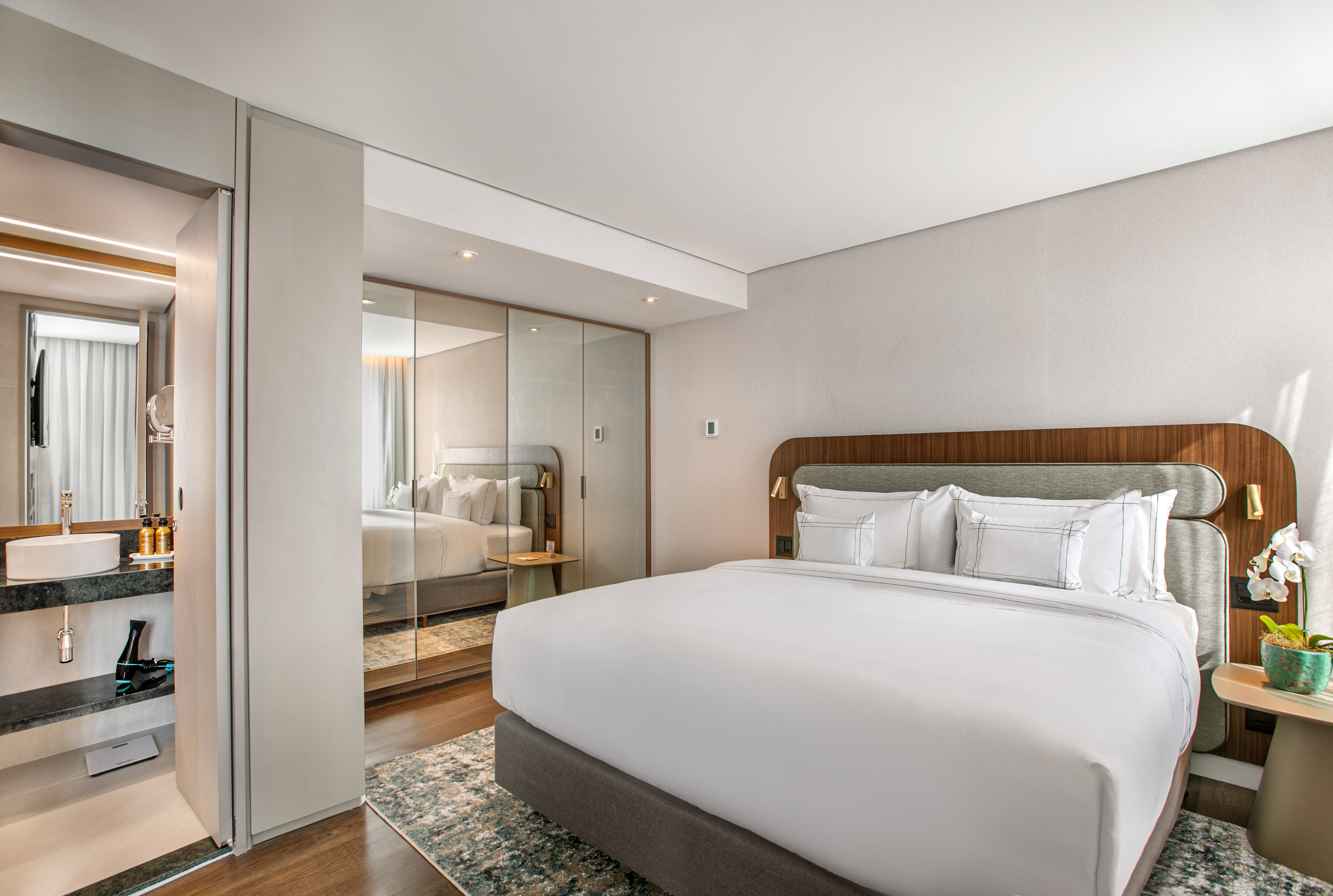 Modern hotel bedroom with king bed, reflective wardrobe, and ensuite vanity.