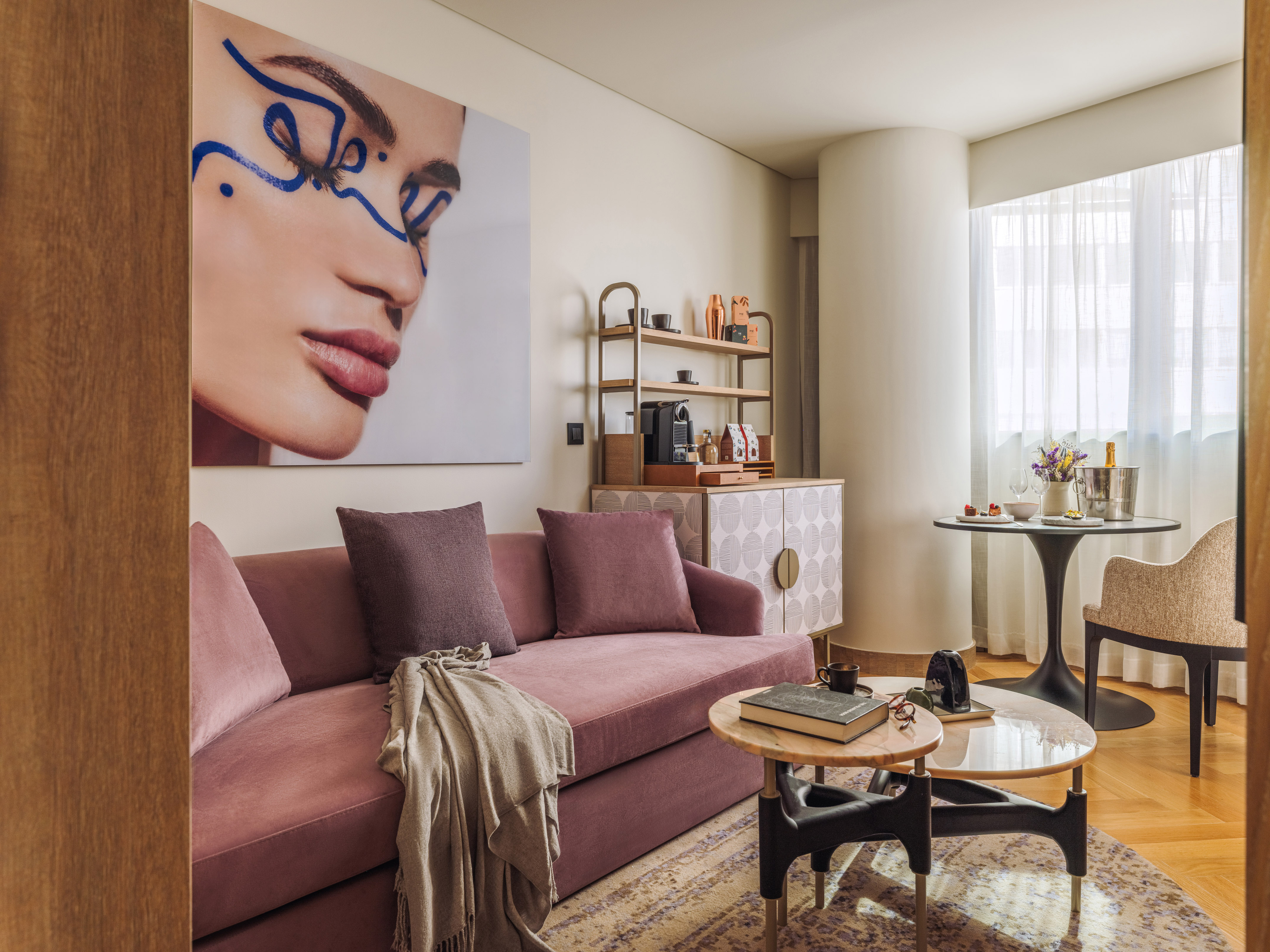Stylish hotel suite with pink sofa, abstract art, modern minibar, dining nook.