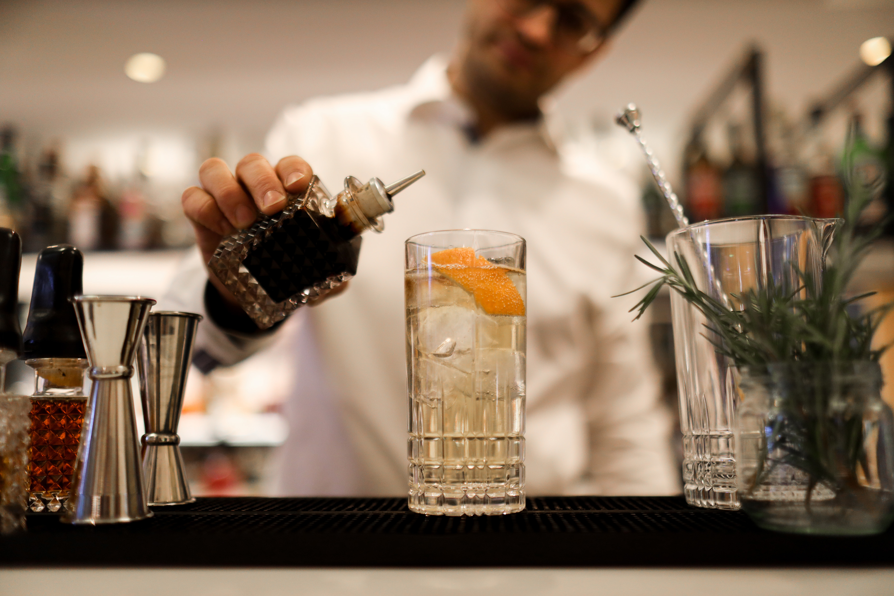 Bartender pouring cocktail from bottle into glass with ice, orange peel garnish.