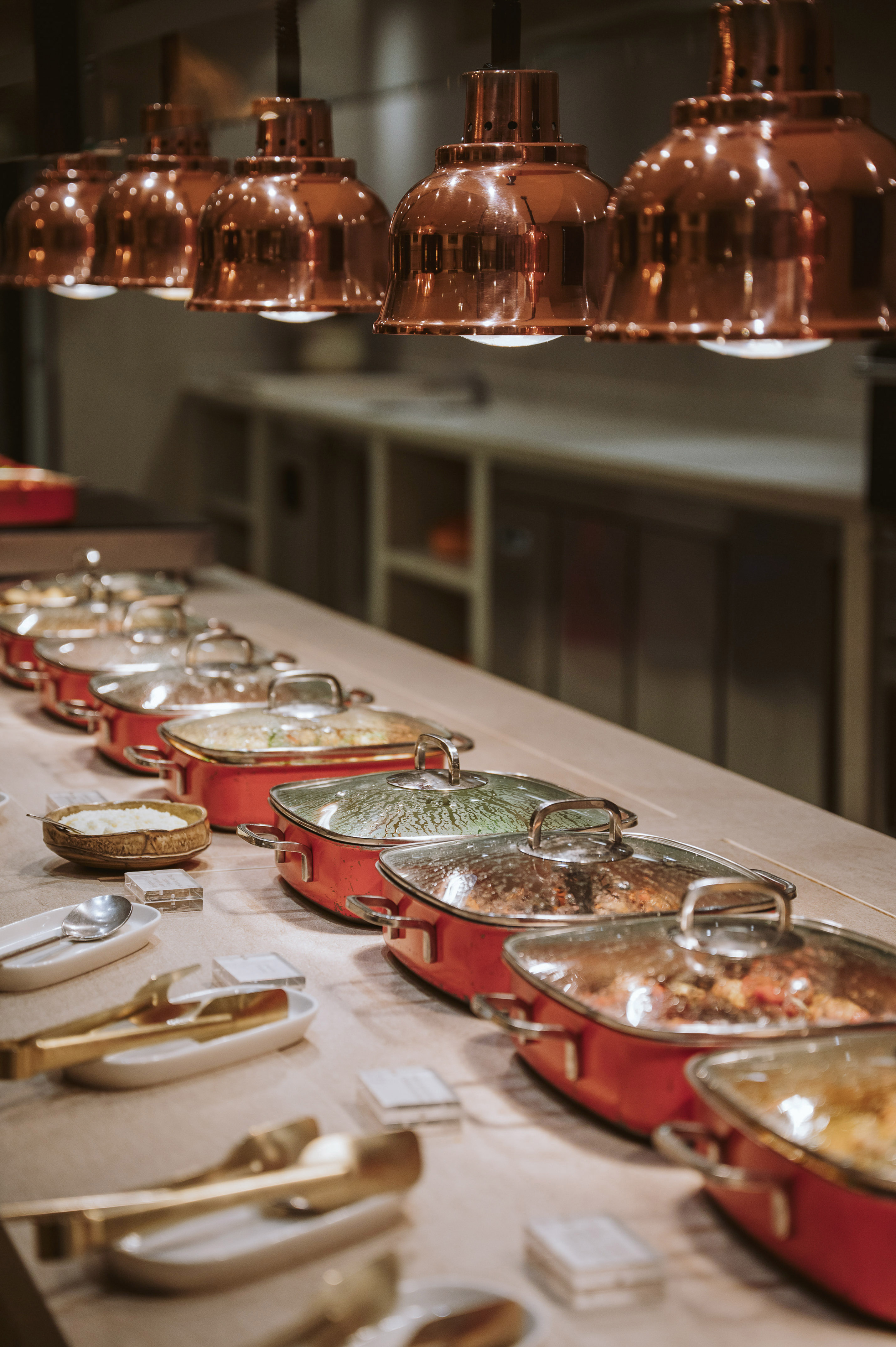 Buffet line with hot food in red serving pans under copper heat lamps.