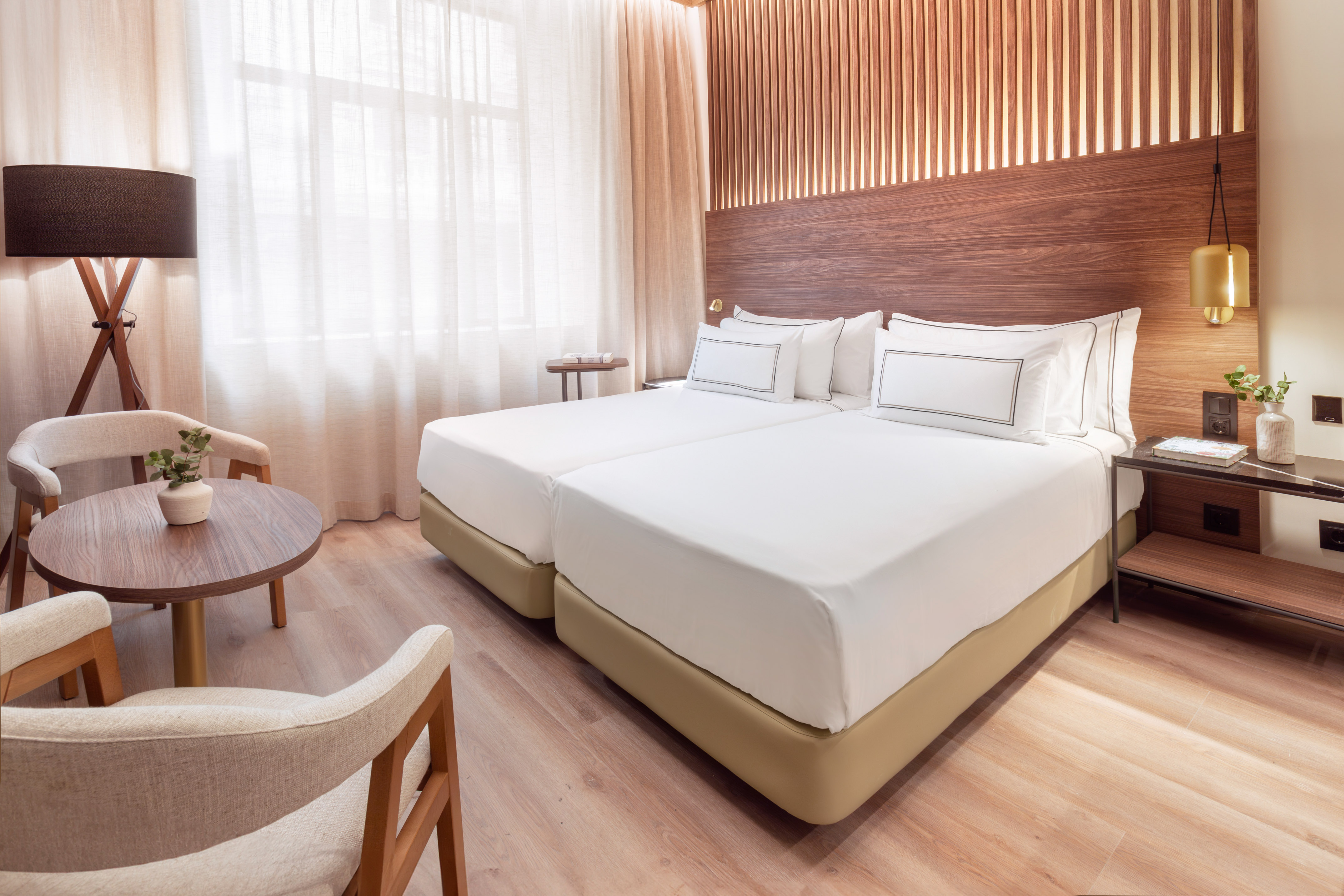 Habitación de hotel moderna con dos camas, cabecero de madera y zona de estar.