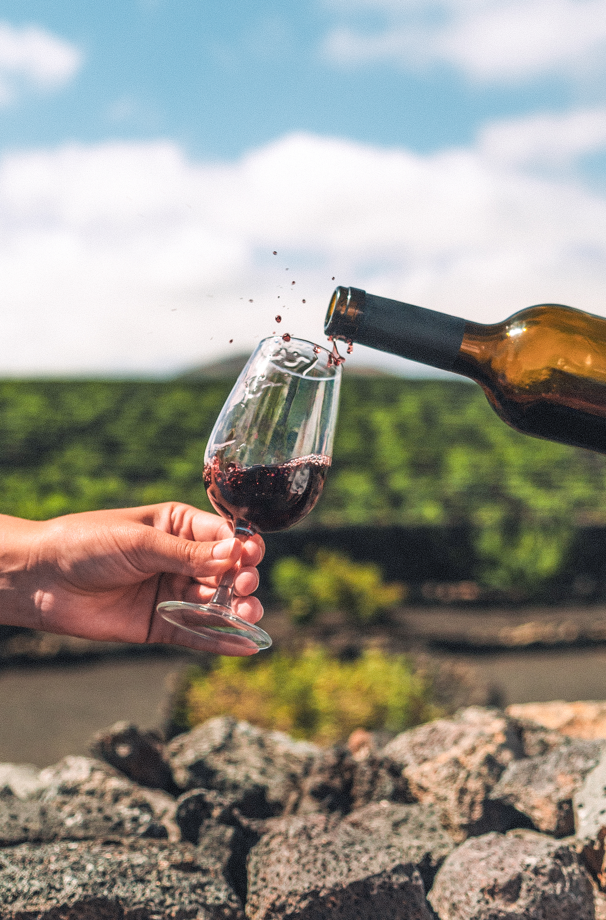 Red wine pouring from bottle into glass with splashes, green vineyard background.