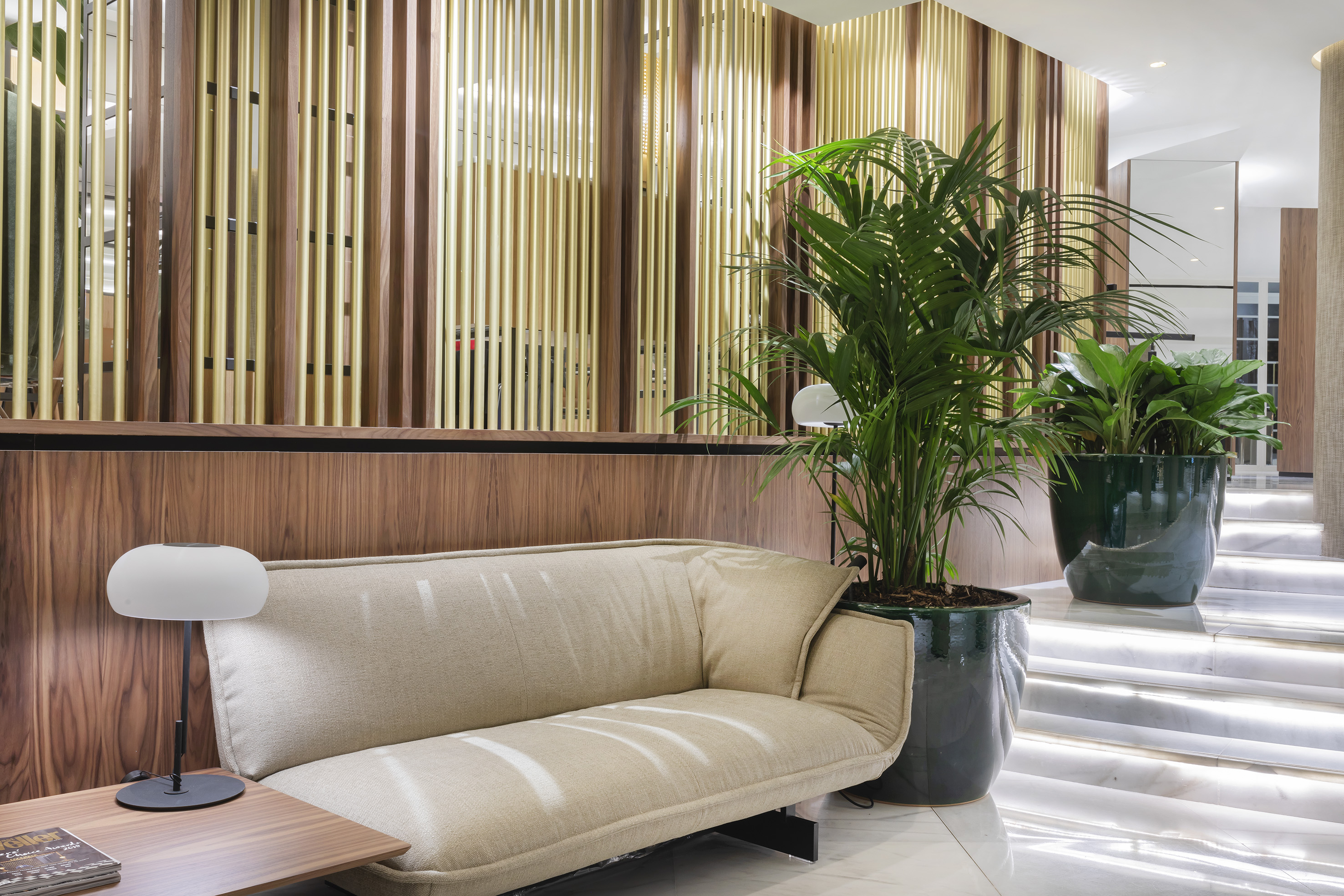 Modern hotel lounge with beige sofa, wood-gold wall, and lush potted plants.