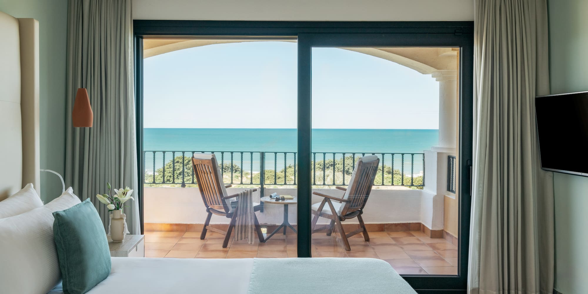 Hotel bedroom with ocean view balcony, chairs, and blue sea.