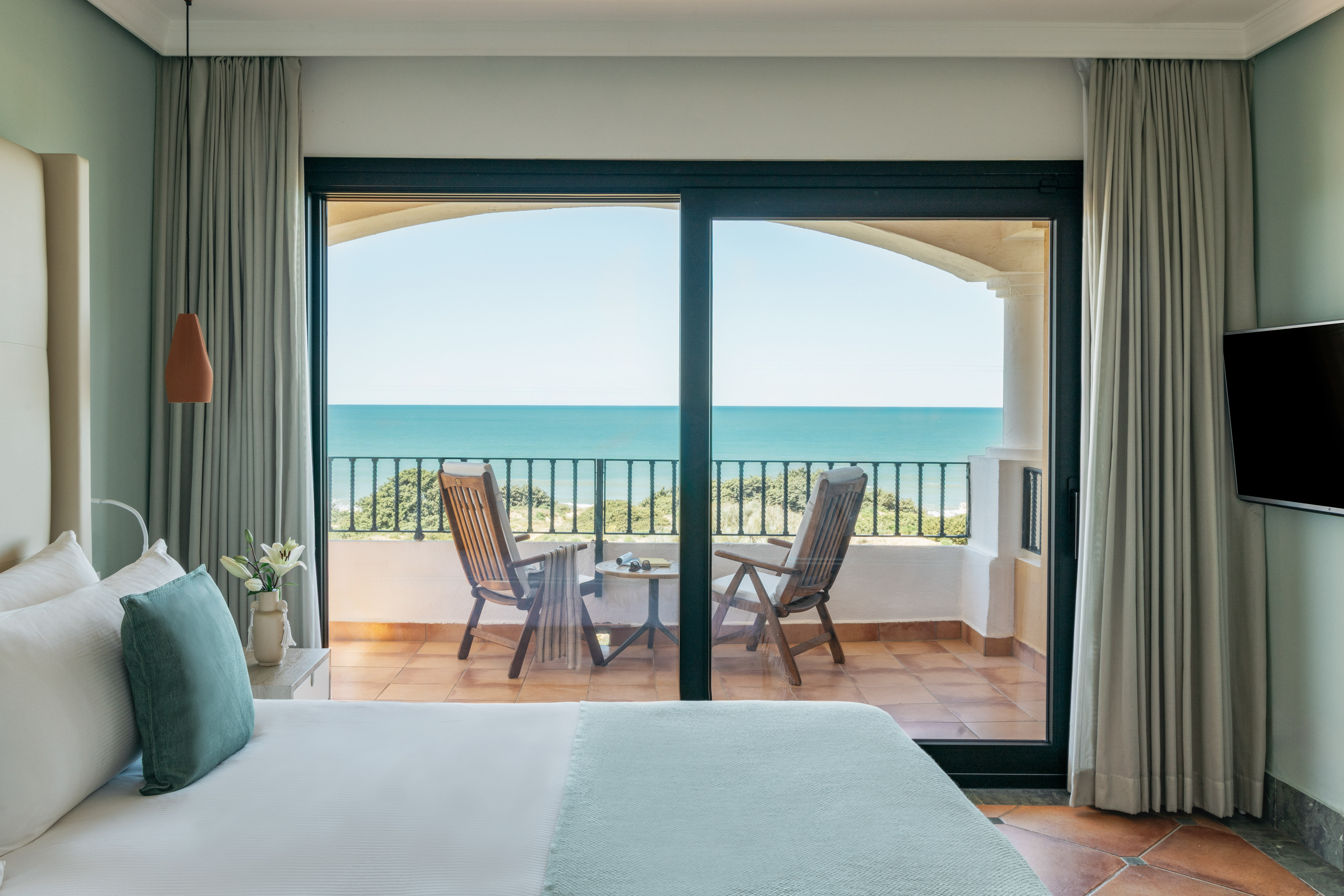 Hotel bedroom with ocean view balcony, chairs, and blue sea.