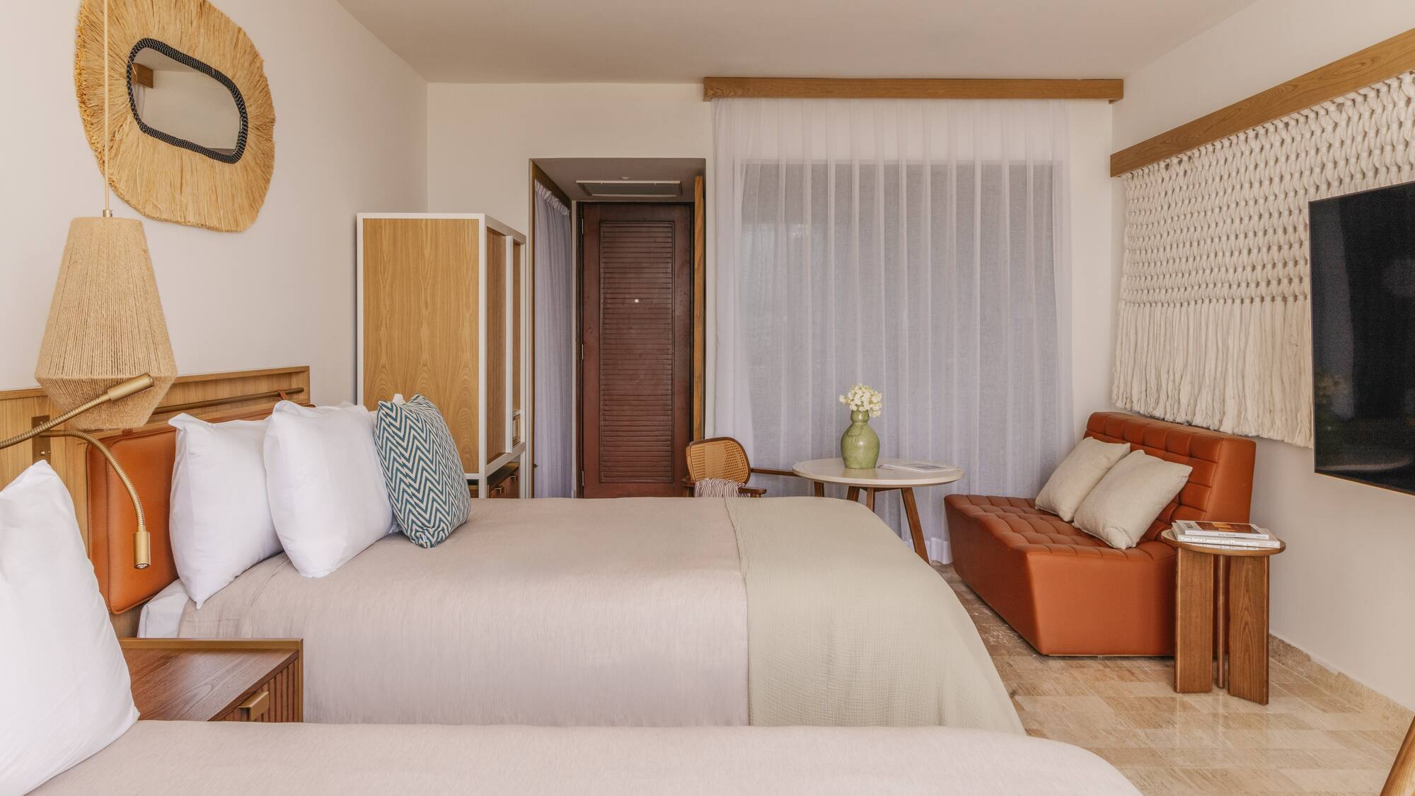 Tropical resort room with two beds, rattan mirror, orange sofa, and woven decor.