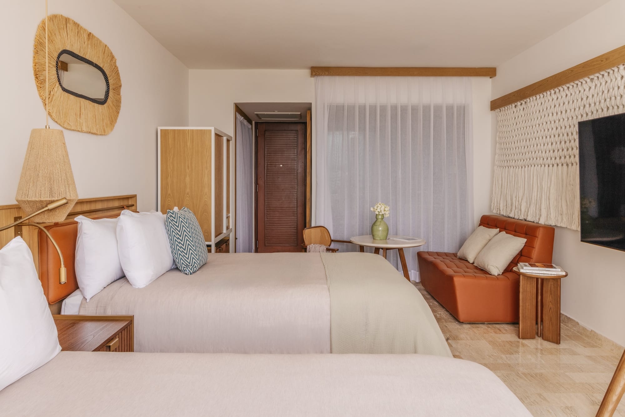 Tropical resort room with two beds, rattan mirror, orange sofa, and woven decor.