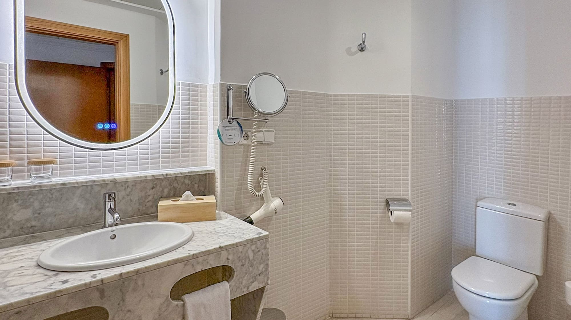 Modern hotel bathroom with marble vanity, lighted mirror, white toilet, and tiled walls.