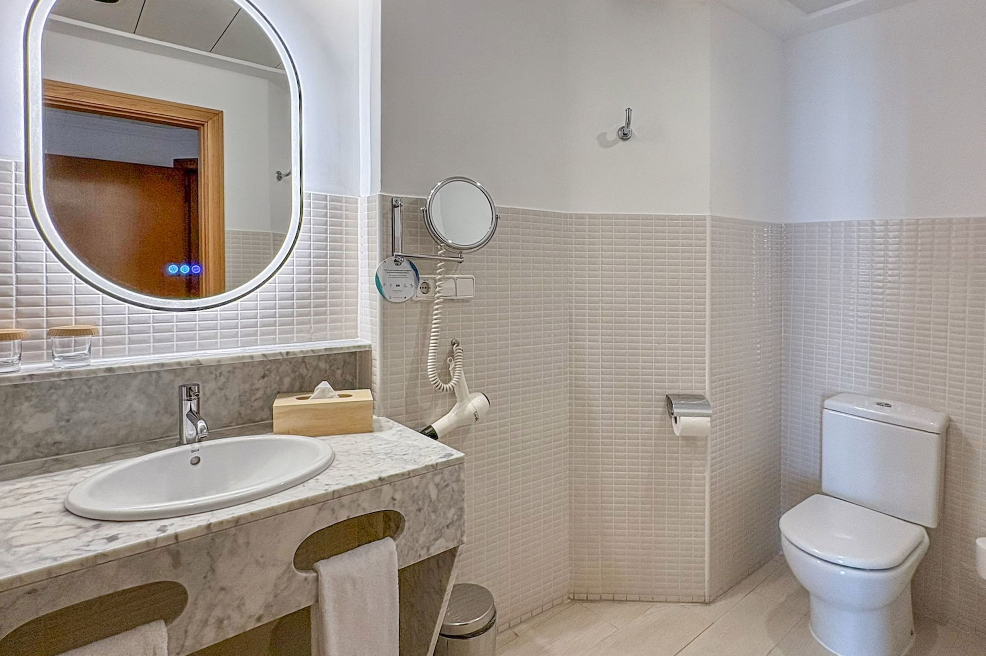 Modern hotel bathroom with marble vanity, lighted mirror, white toilet, and tiled walls.