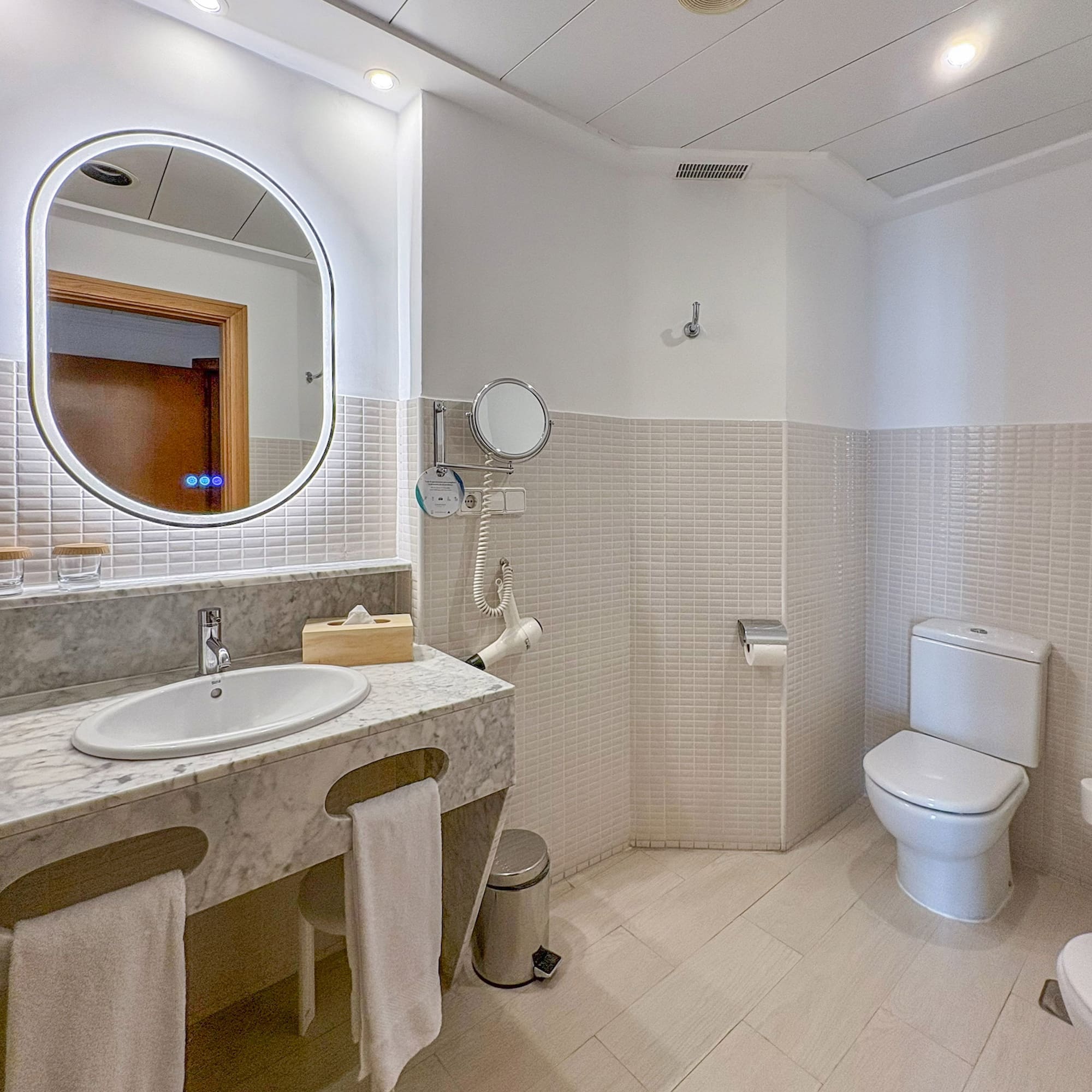 Modern hotel bathroom with marble vanity, lighted mirror, white toilet, and tiled walls.