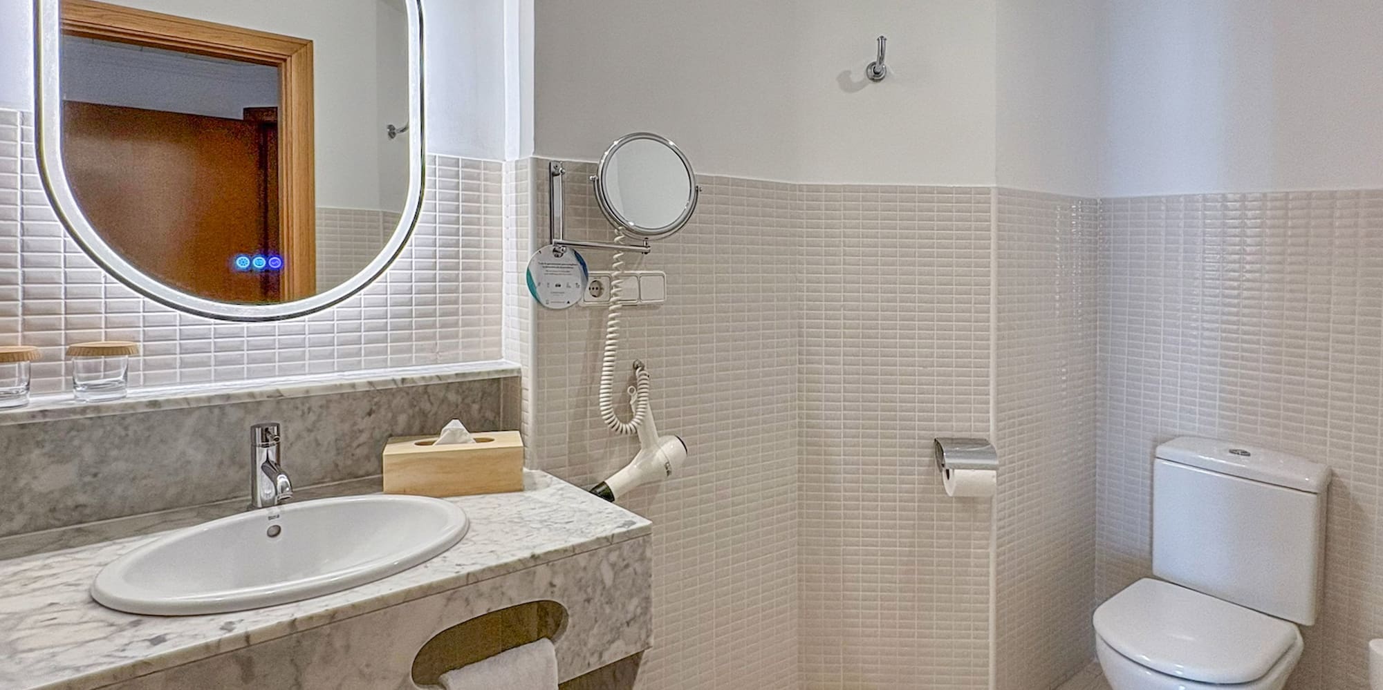 Modern hotel bathroom with marble vanity, lighted mirror, white toilet, and tiled walls.