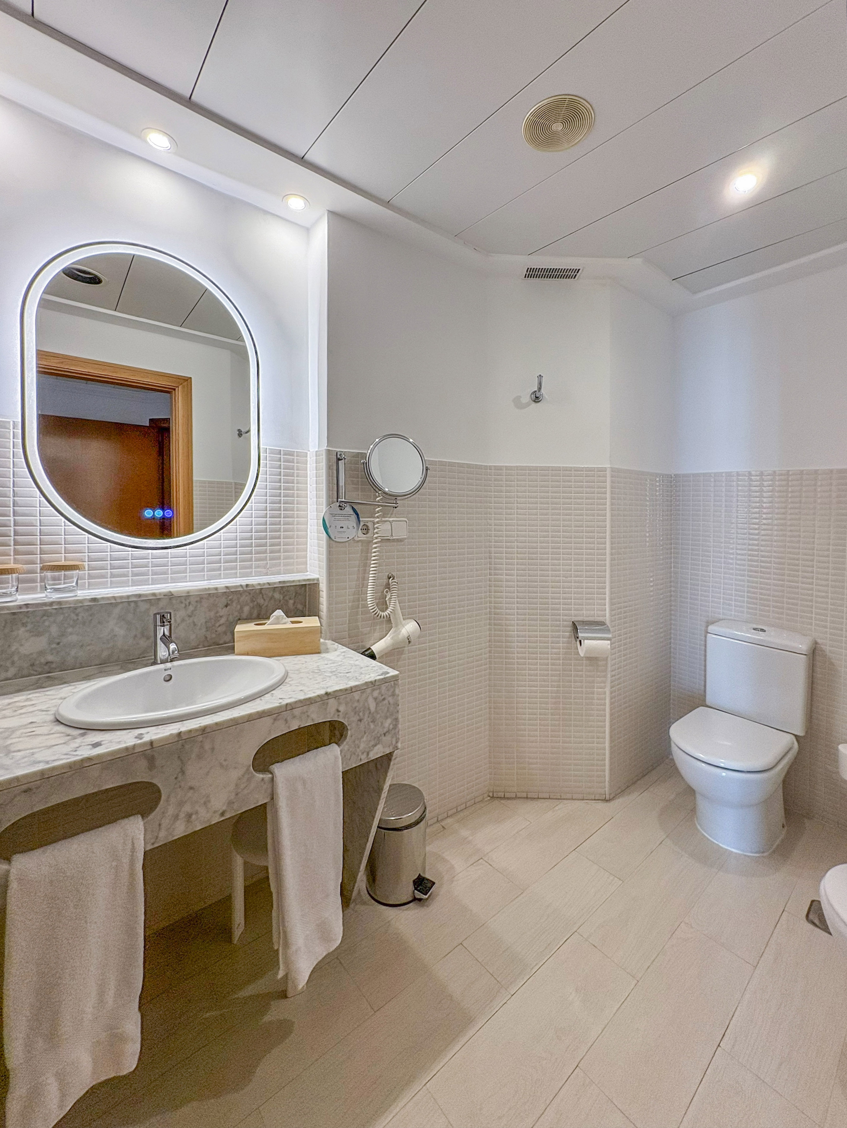 Modern hotel bathroom with marble vanity, lighted mirror, white toilet, and tiled walls.