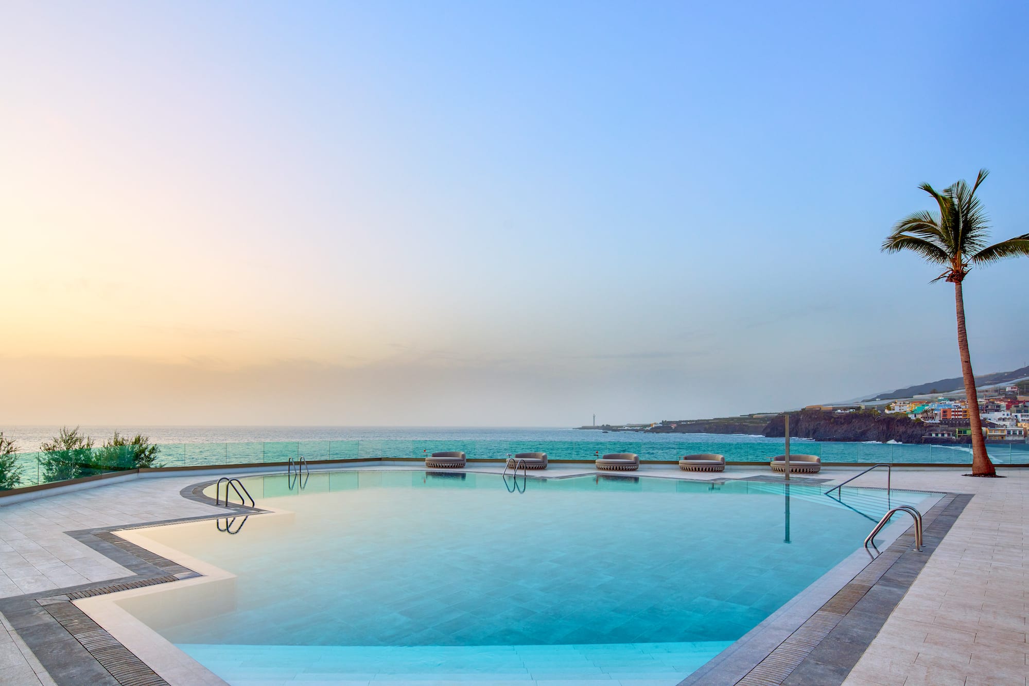 Infinity pool overlooking ocean at sunset, with a palm tree and distant coast.