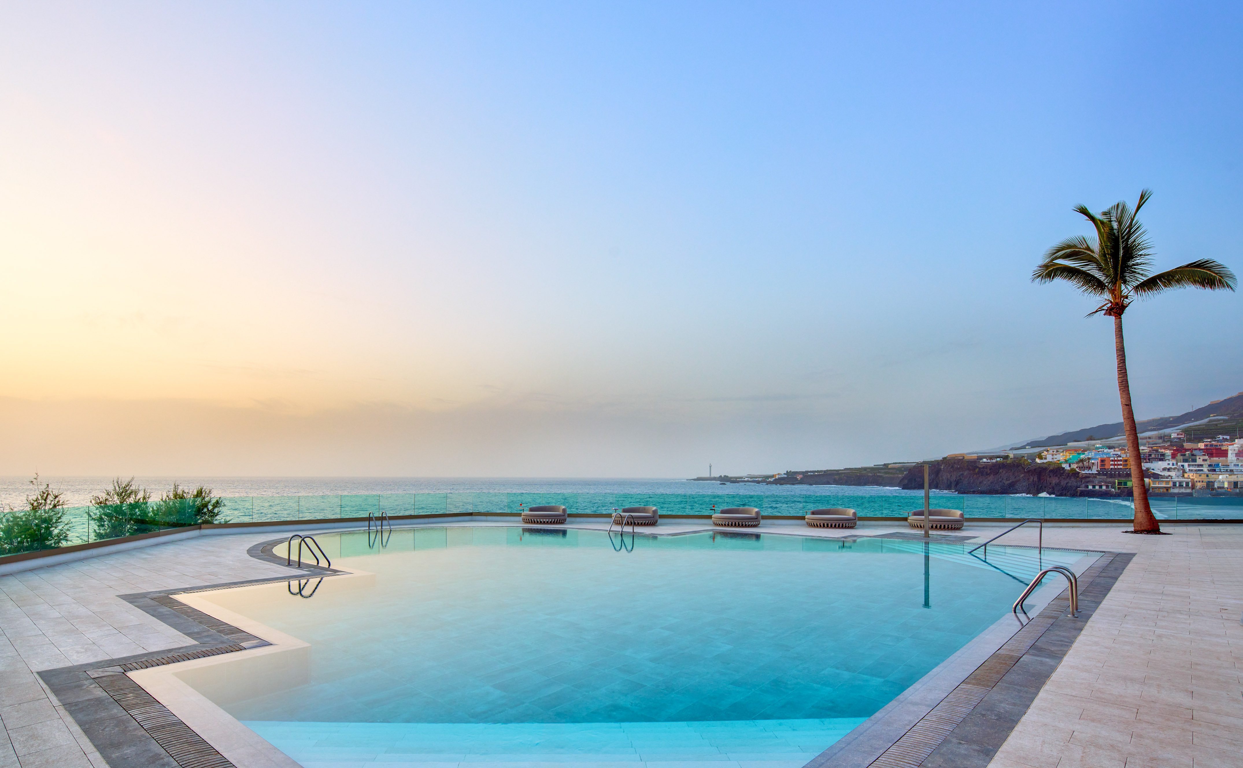 Infinity pool overlooking ocean at sunset, with a palm tree and distant coast.