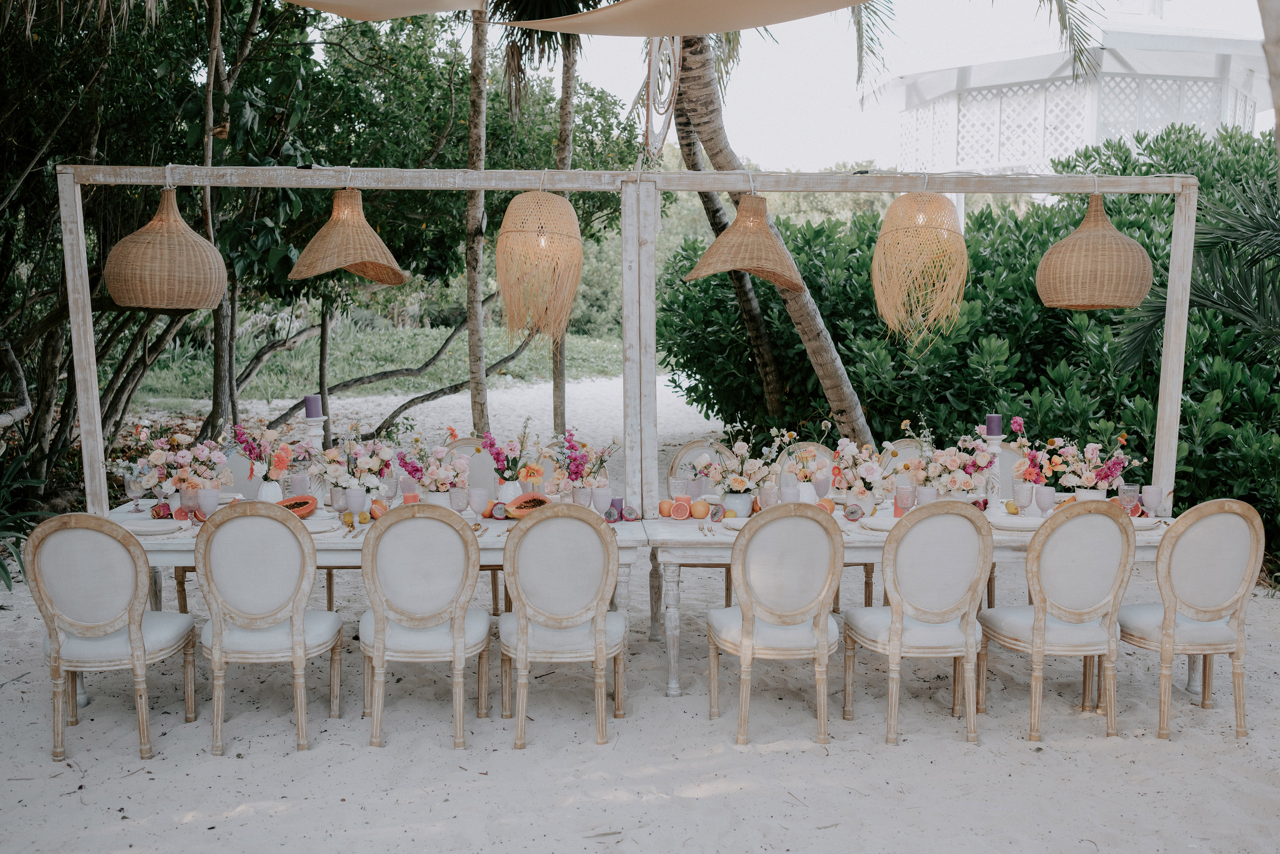 Elegant outdoor dining table on beach, with flowers, fruit, and woven lights.
