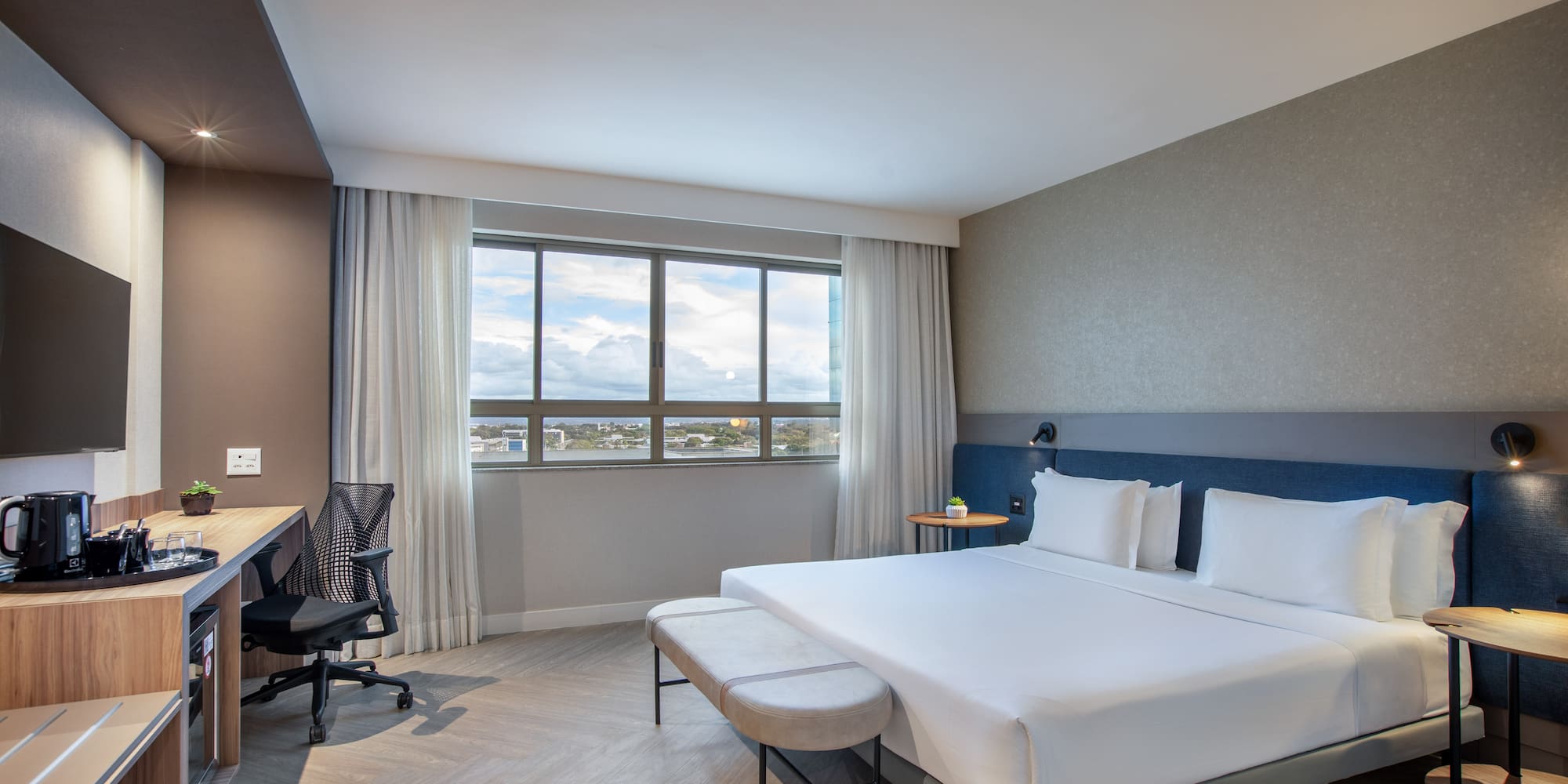 Modern hotel room with queen bed, workspace, and city view window.