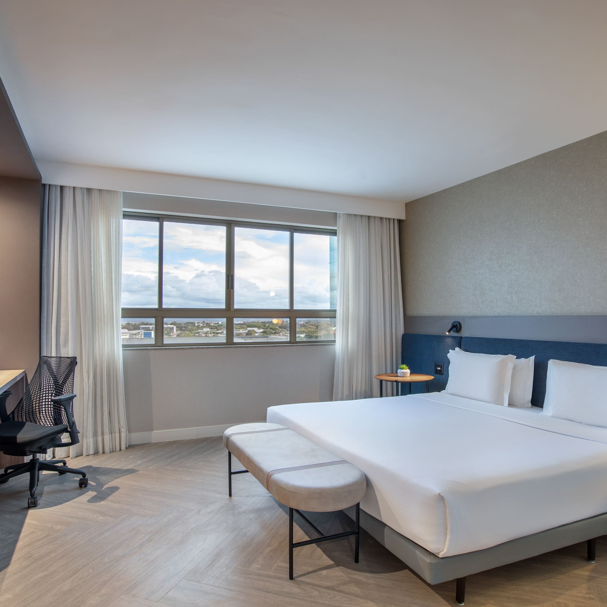 Modern hotel room with queen bed, workspace, and city view window.