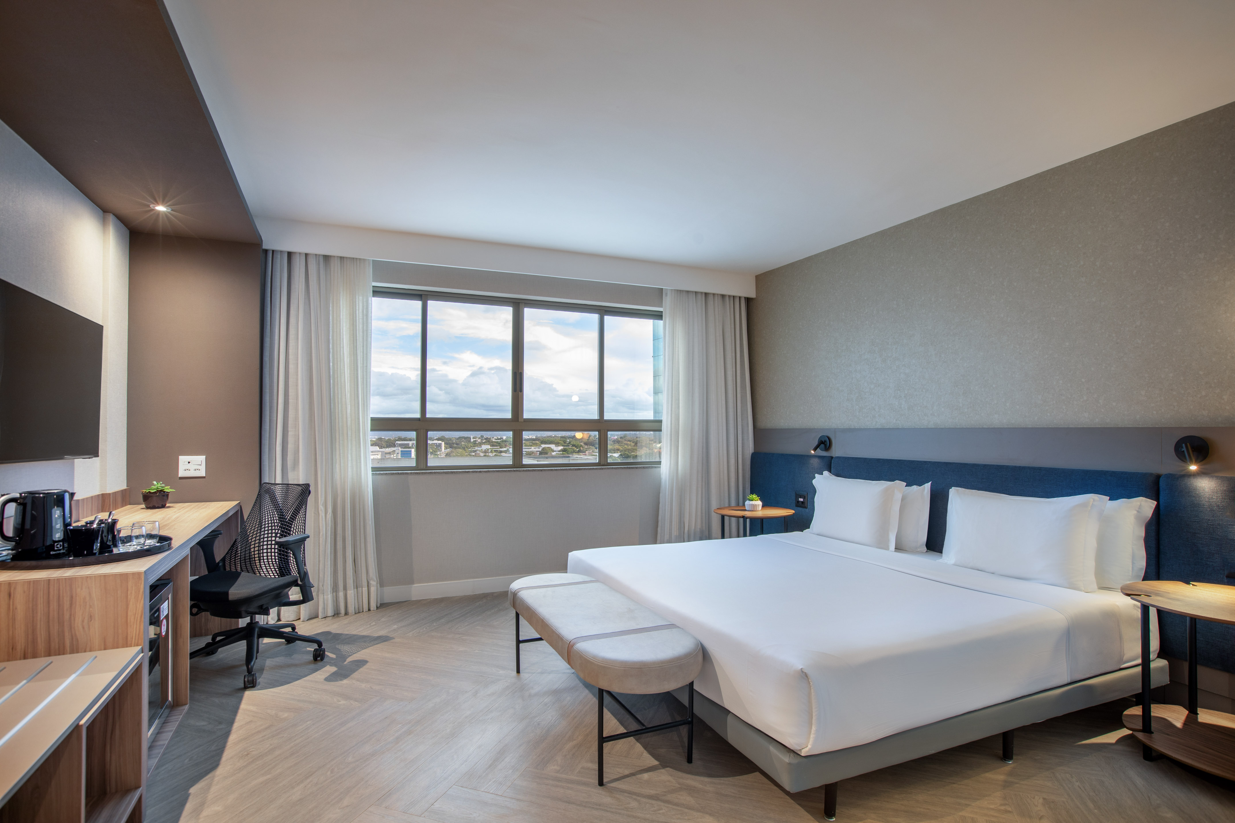 Modern hotel room with queen bed, workspace, and city view window.