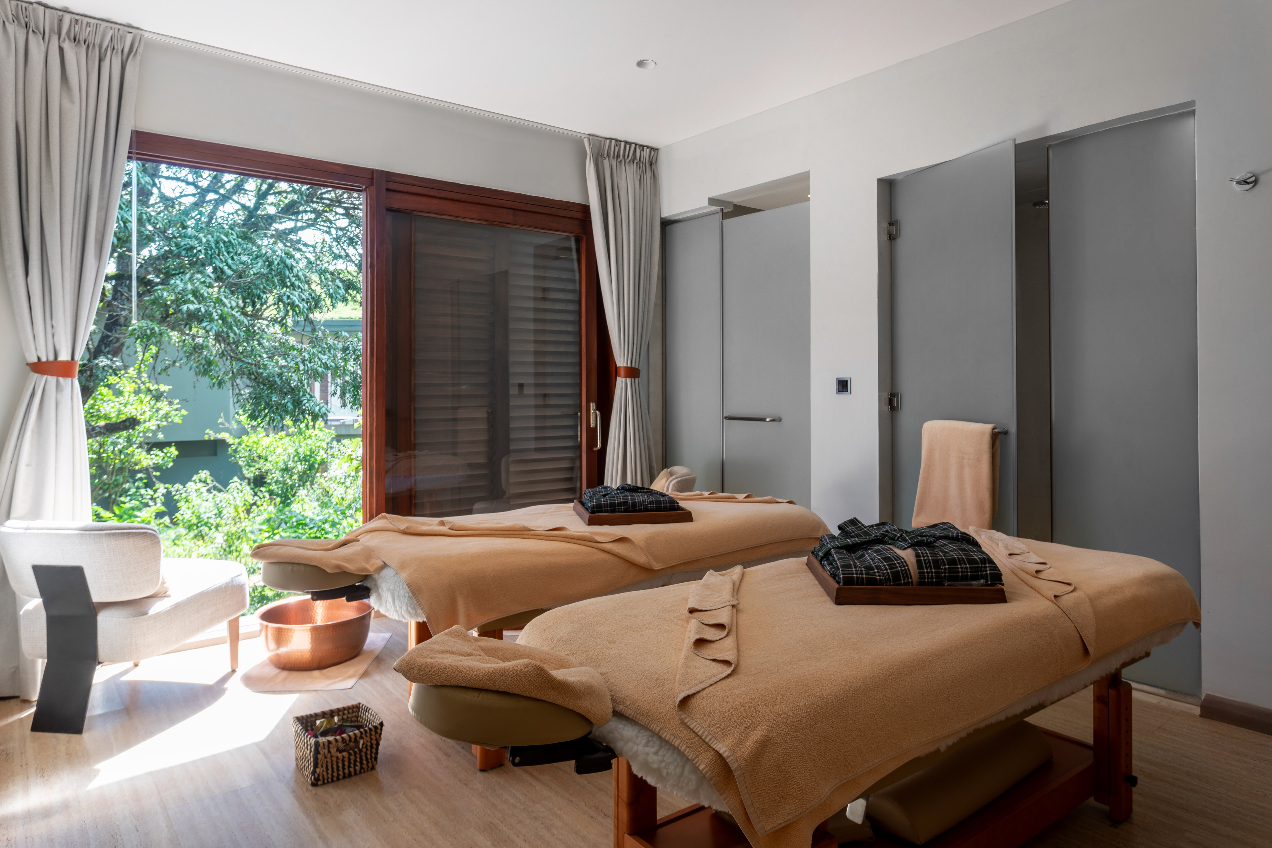 Modern spa room with two massage tables, robes, and a large window with greenery.