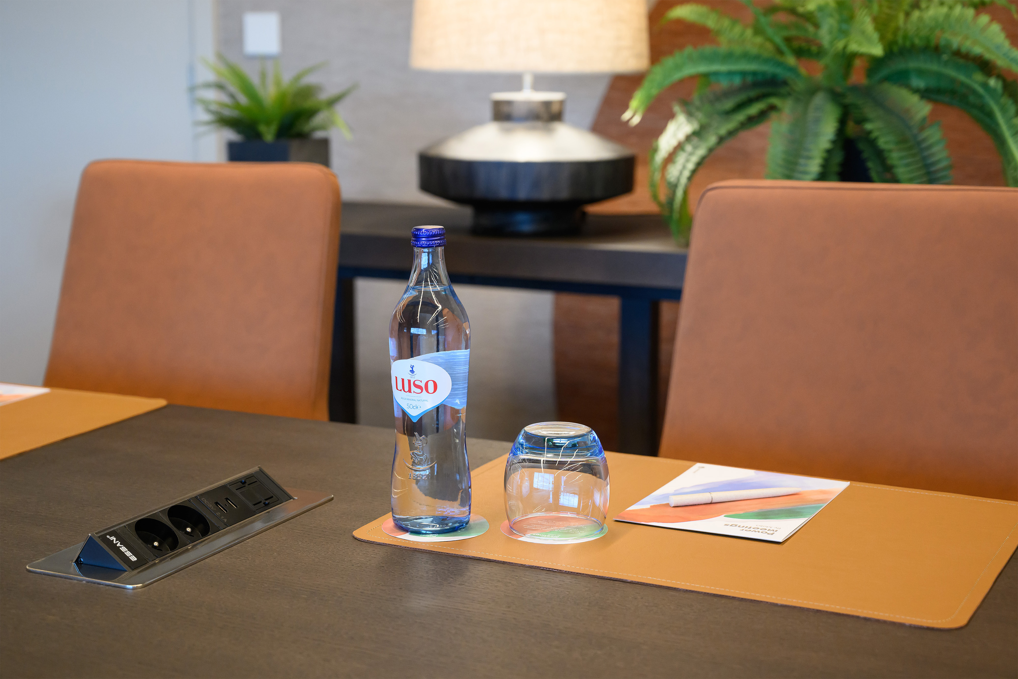 Meeting table with Luso water bottle, glass, power outlet, and tan leather desk pad.